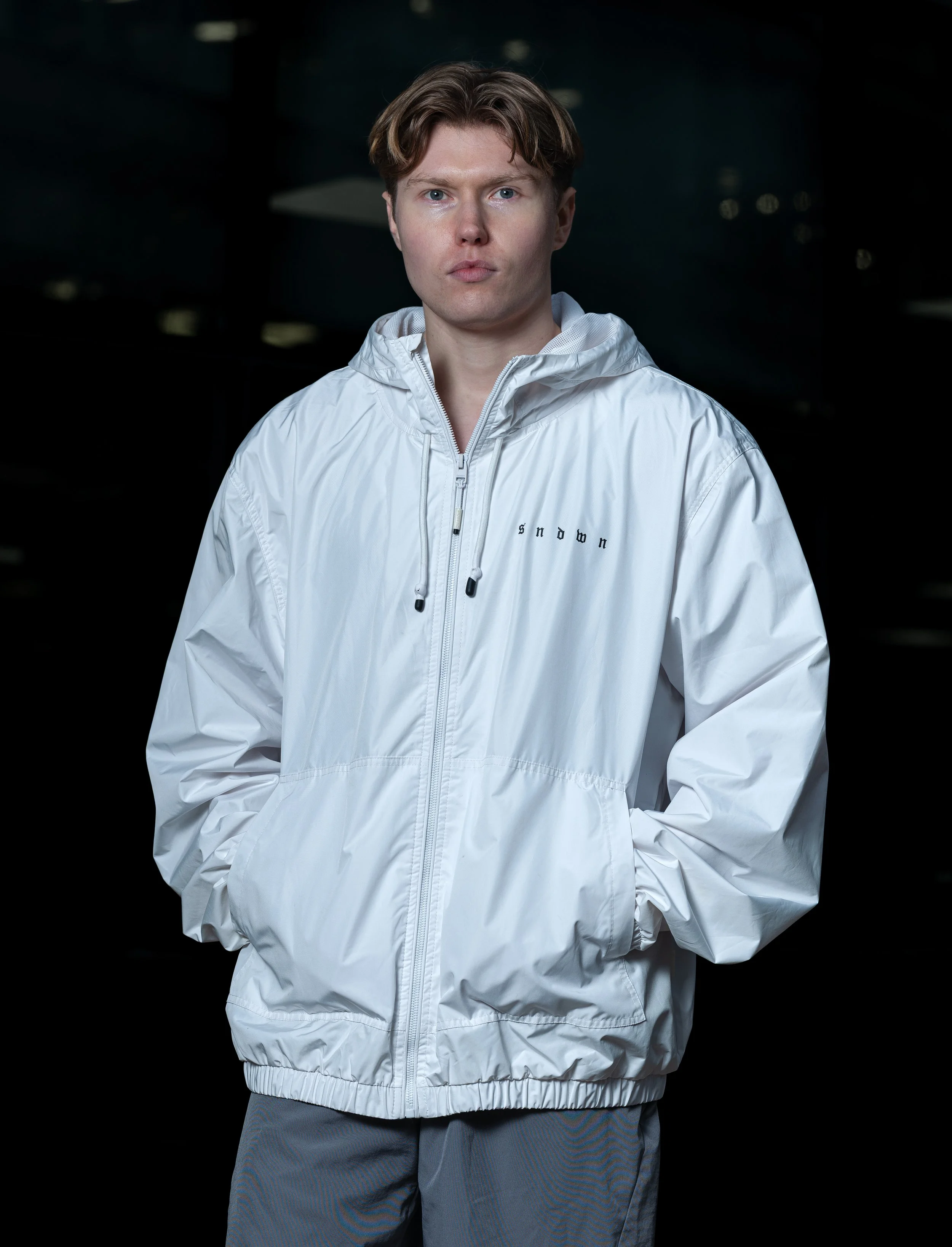 A young man with short brown hair wearing a white windbreaker with the word 'snow' embroidered on the chest, standing against a dark background.