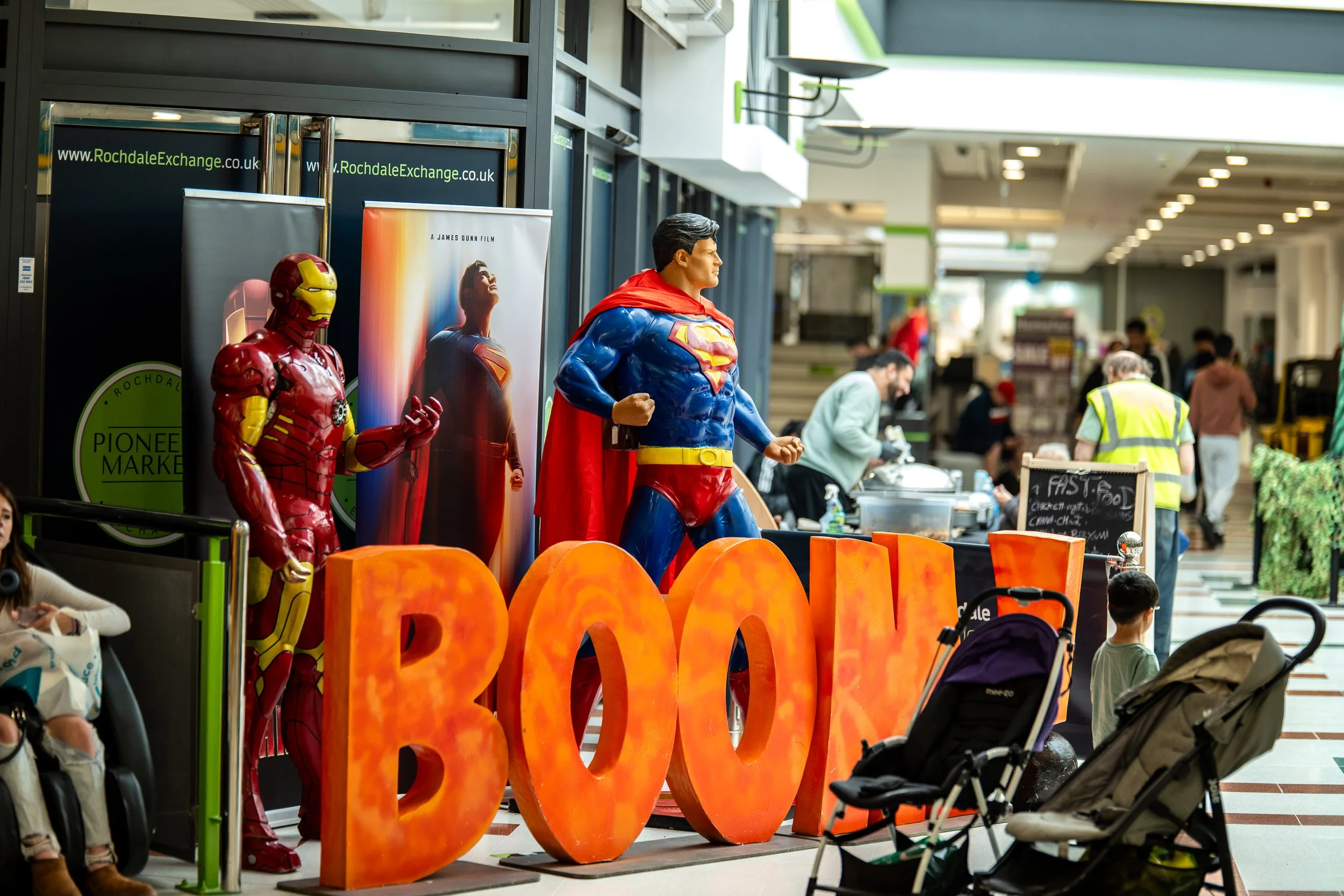 Display in a shopping mall featuring life-sized superhero statues of Iron Man, Supergirl, and Superman, with large illuminated letters spelling 'BOO' in front, and people shopping and sitting around.