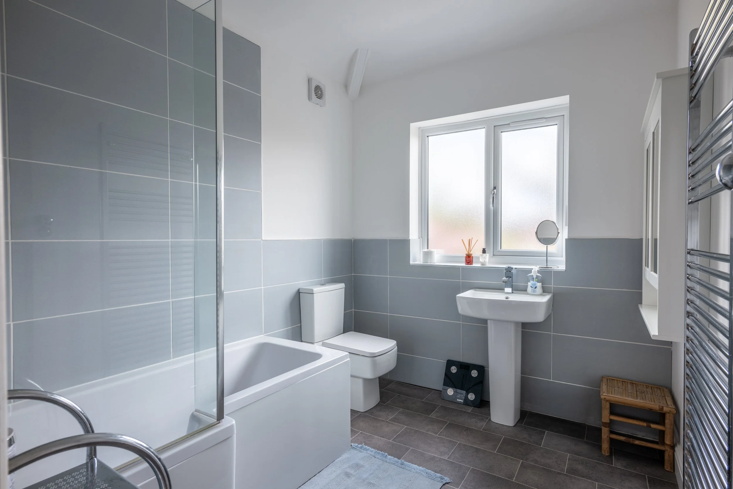 Modern bathroom with gray tiled walls, white toilet, white bathtub, pedestal sink, frosted window with small decorative items, electric towel warmer, and wooden stool.