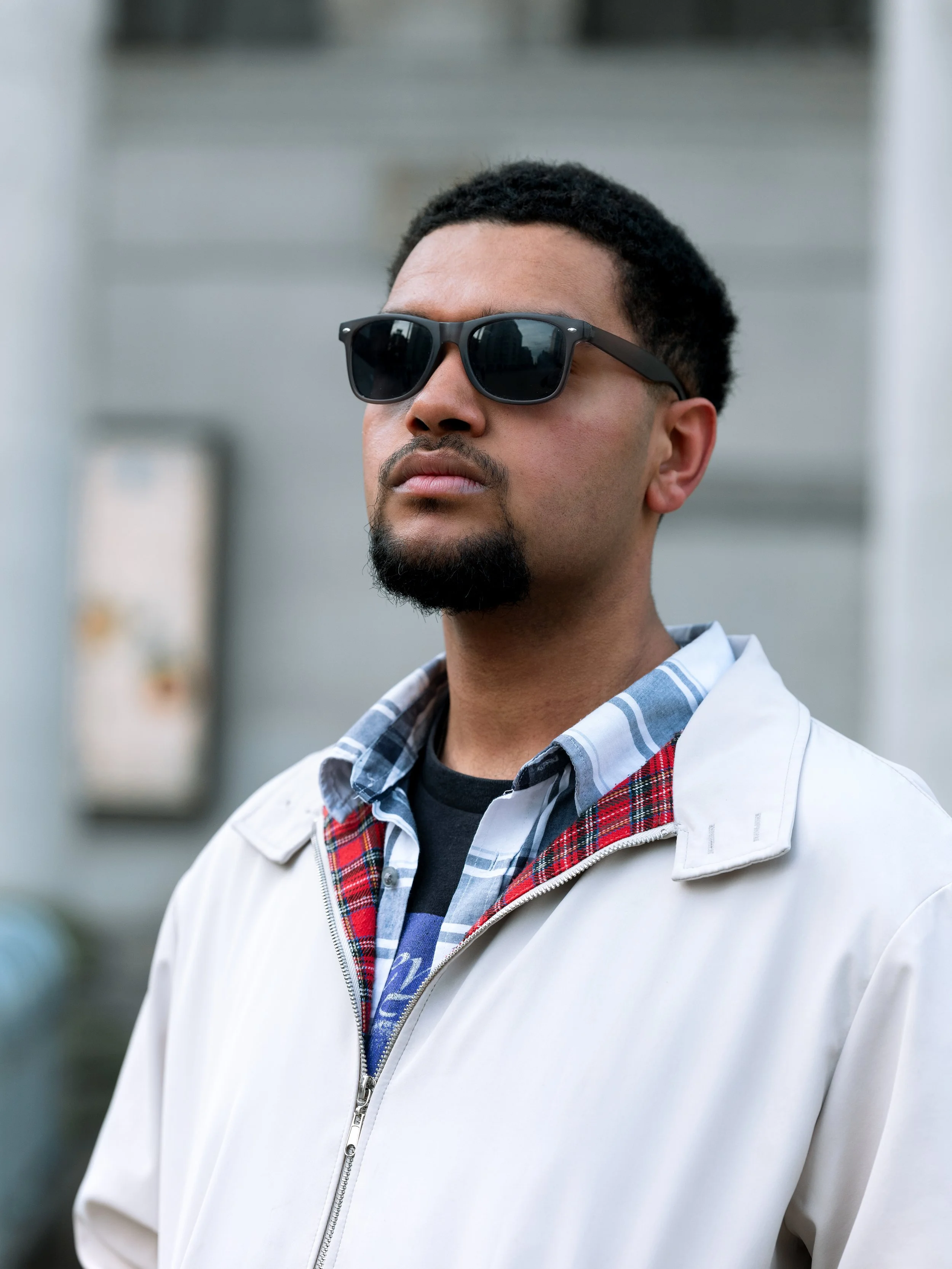 A man wearing sunglasses, a plaid shirt, and a white jacket standing outdoors with a neutral expression.
