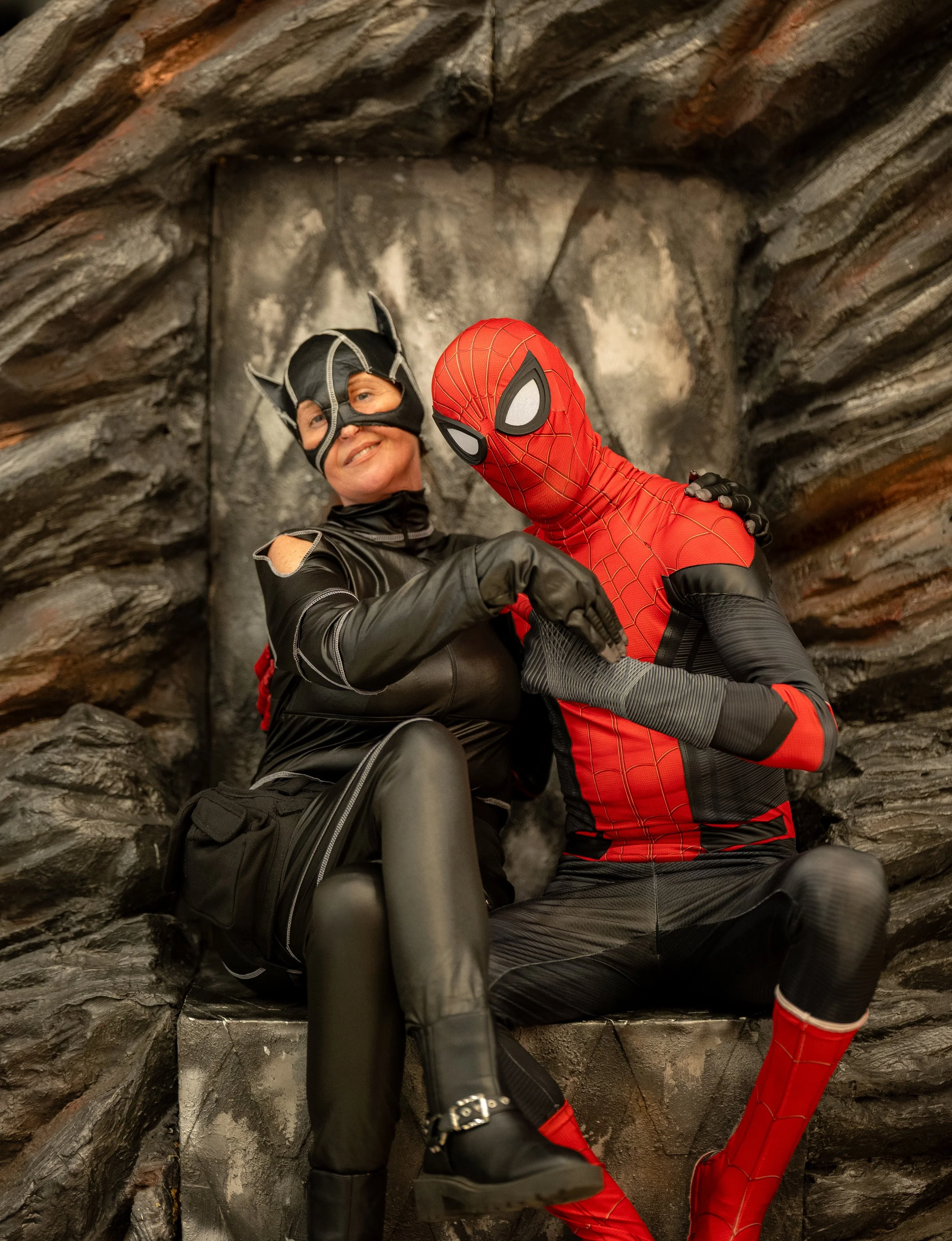 Two people in superhero costumes, Batman and Spider-Man, sitting together on a rock formation. Batman is smiling, and Spider-Man has his arm around Batman.