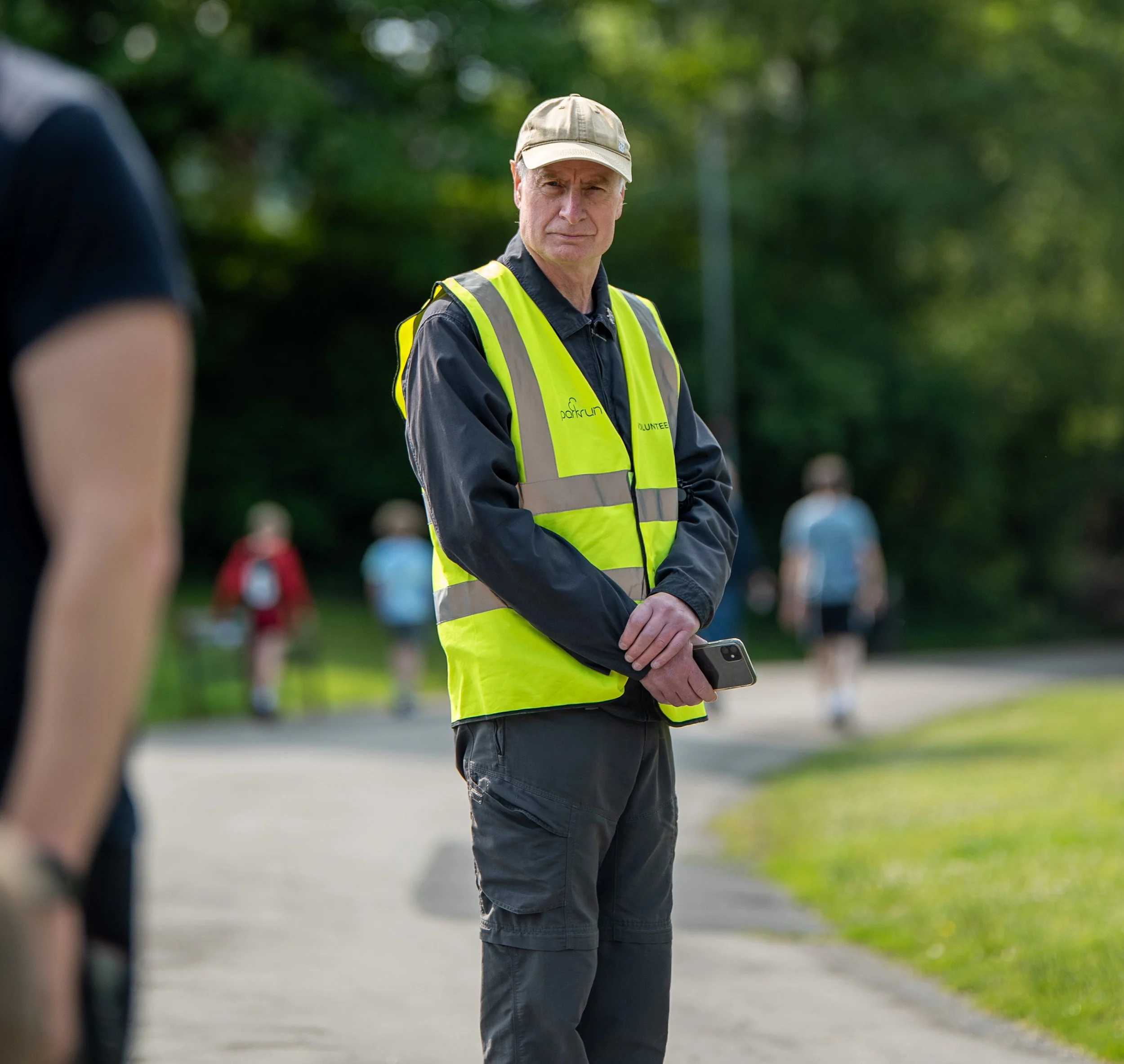 A man wearing a yellow safety vest and a beige cap stands outdoors on a sidewalk, holding a smartphone, with a grassy area and people blurred in the background.