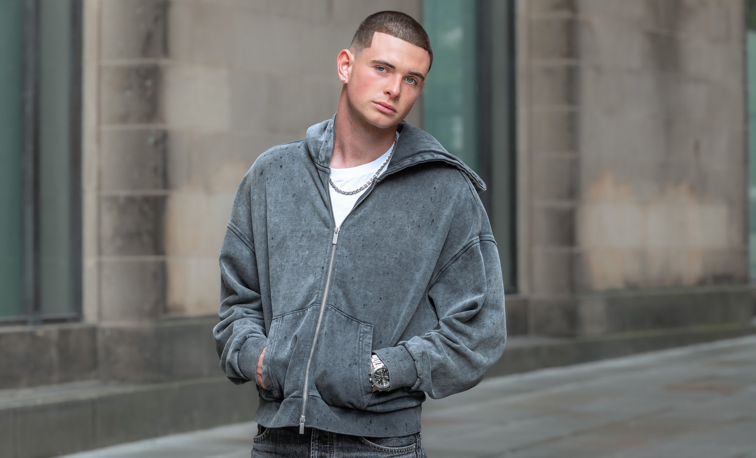 A young man with short hair and light skin wearing a gray zip-up hoodie, white t-shirt, and jewelry, standing outdoors in front of a modern building.
