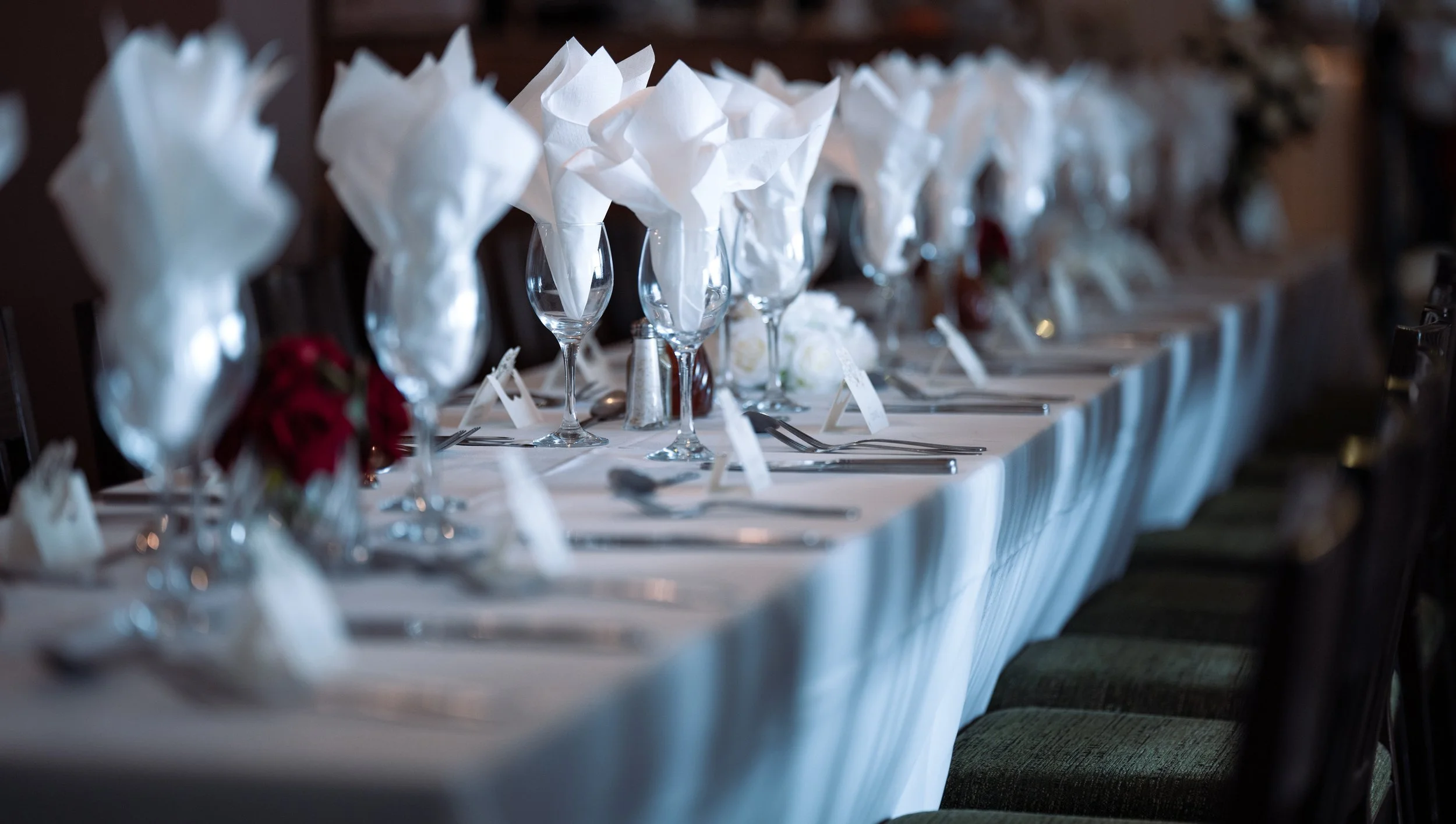 A long banquet table set for a formal event, with white tablecloth, wine glasses with white napkins folded inside, silverware arranged neatly, and small floral arrangements along the center.