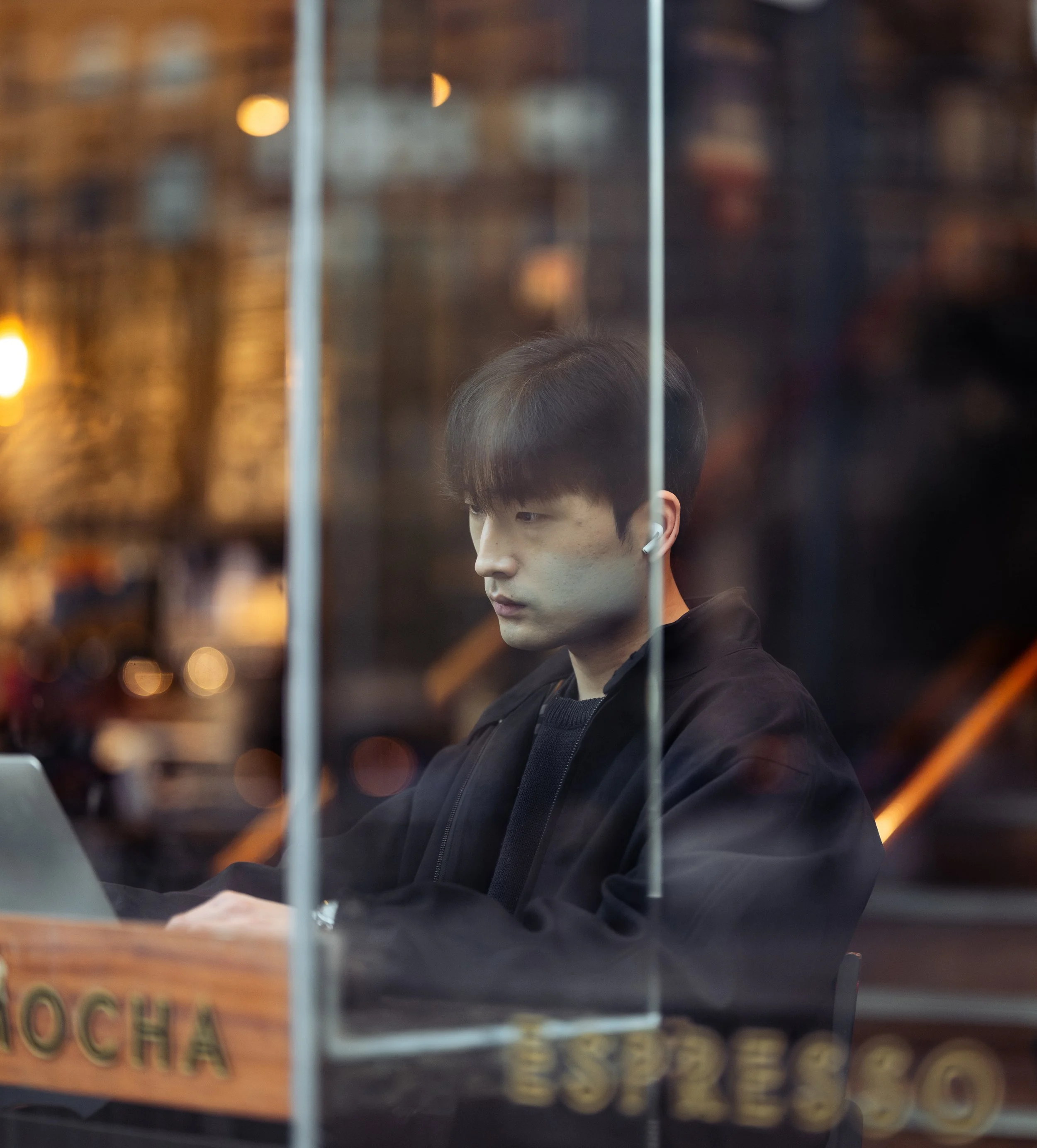 Young man with dark hair wearing a black jacket, looking at a laptop inside a cafe with a glass partition, blurred warm lighting and cafe decor in the background.