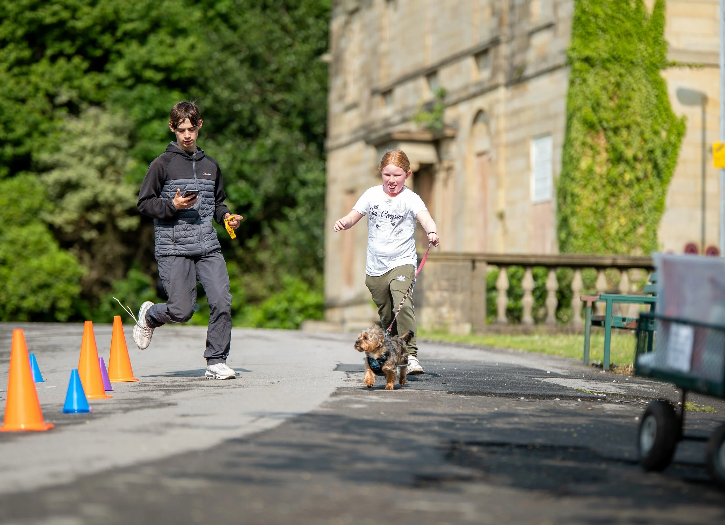 A young girl running with a small dog on a leash on a pathway, while a young boy stands nearby looking at his phone. There are colored cones set up on the pathway. The background features green trees and a stone building with ivy.