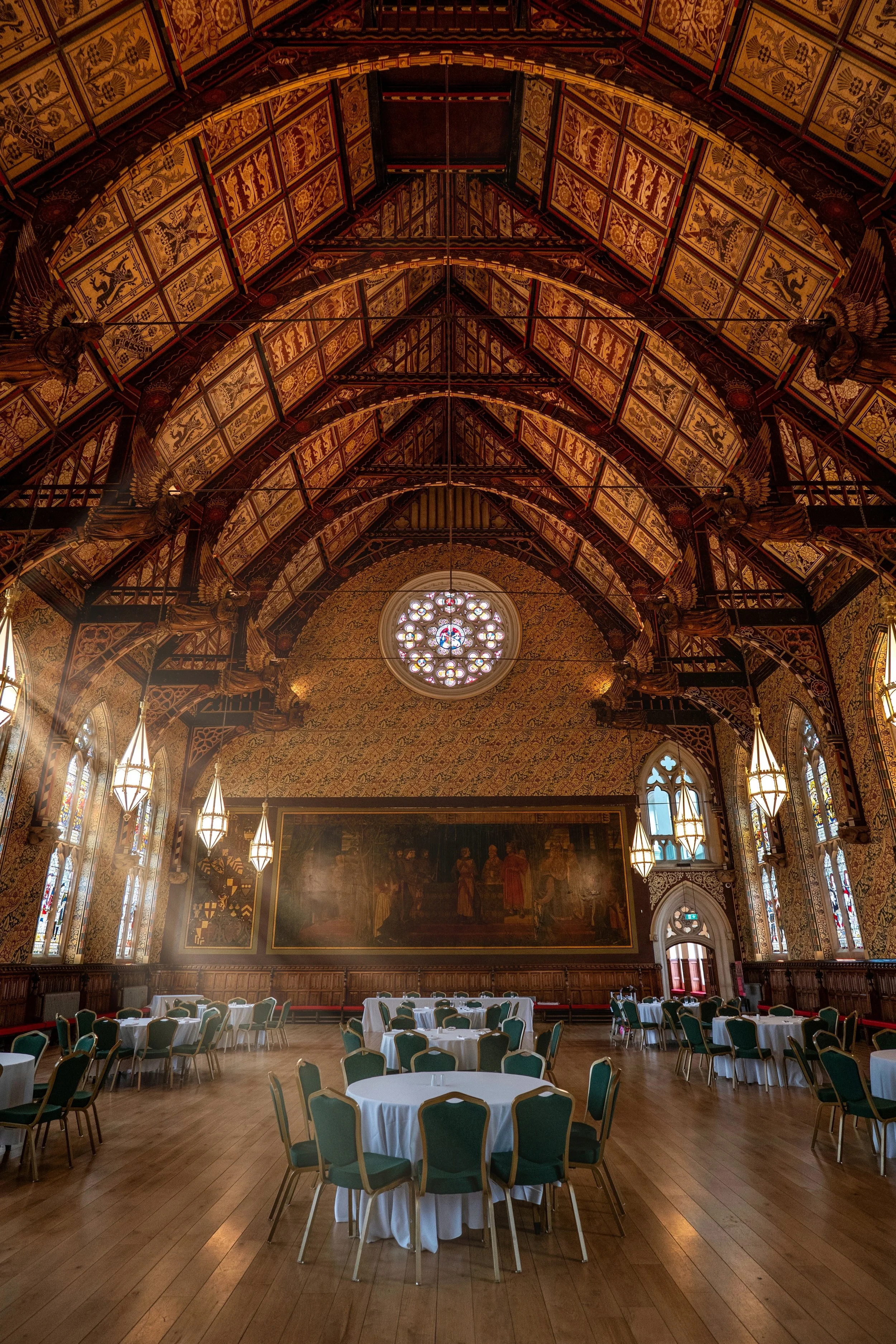 Elegant banquet hall with high, intricately decorated vaulted ceiling, stained glass windows, and round tables with white tablecloths and green chairs.