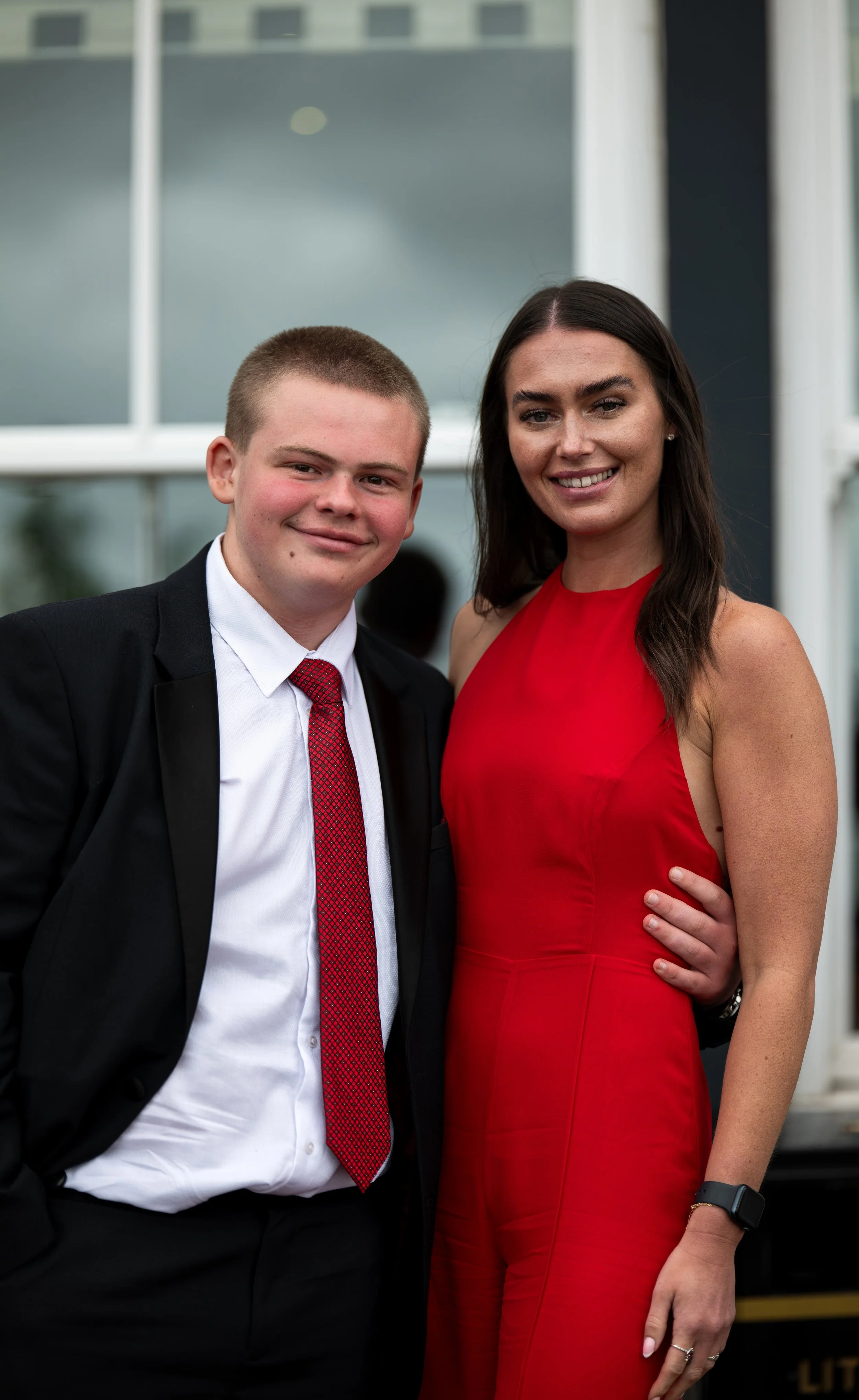 A young man in a black tuxedo with a red tie and a woman in a red dress posing together at a formal event, smiling at the camera.