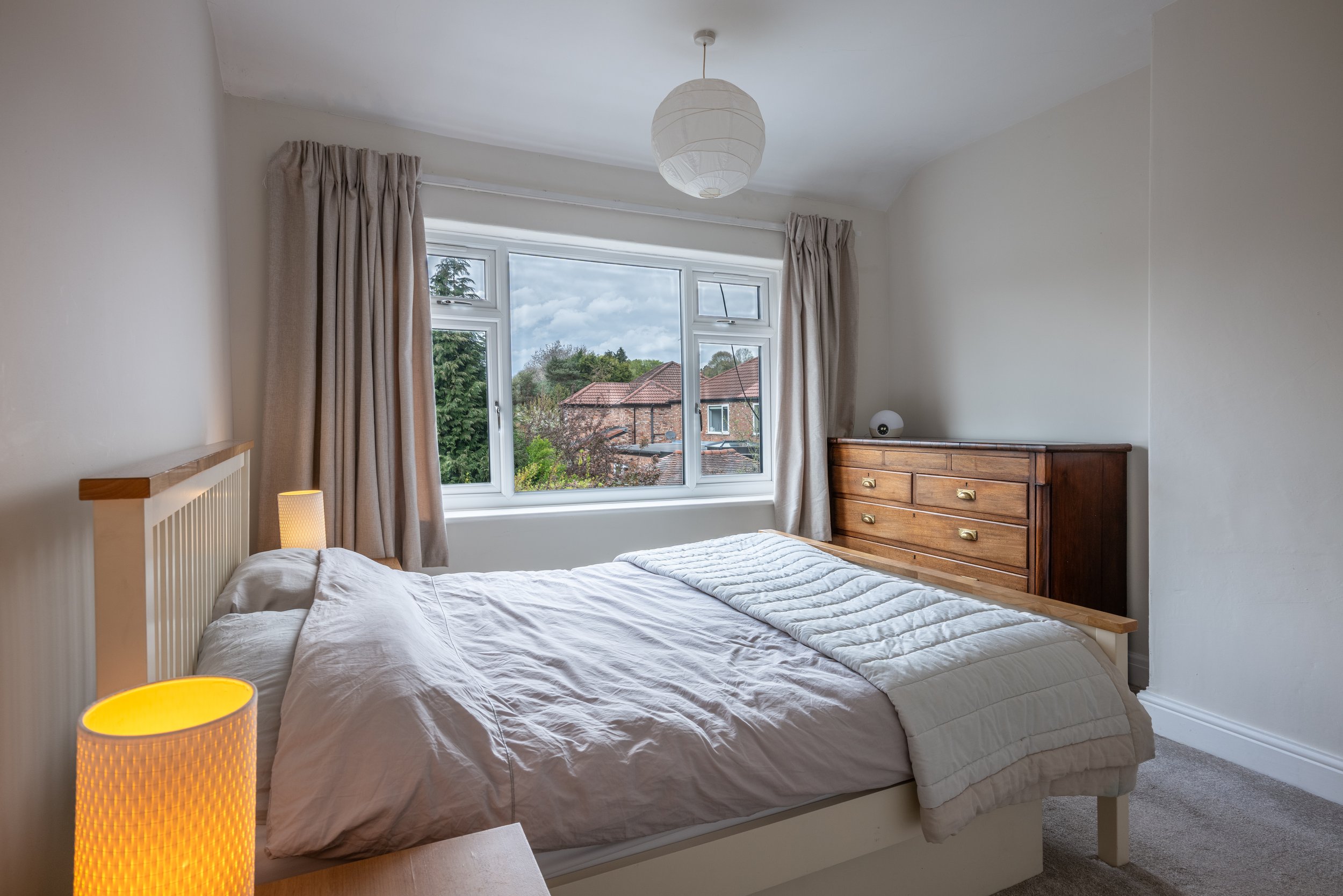 Bedroom with a large window, white bed, beige curtains, a wooden dresser, and lamps.