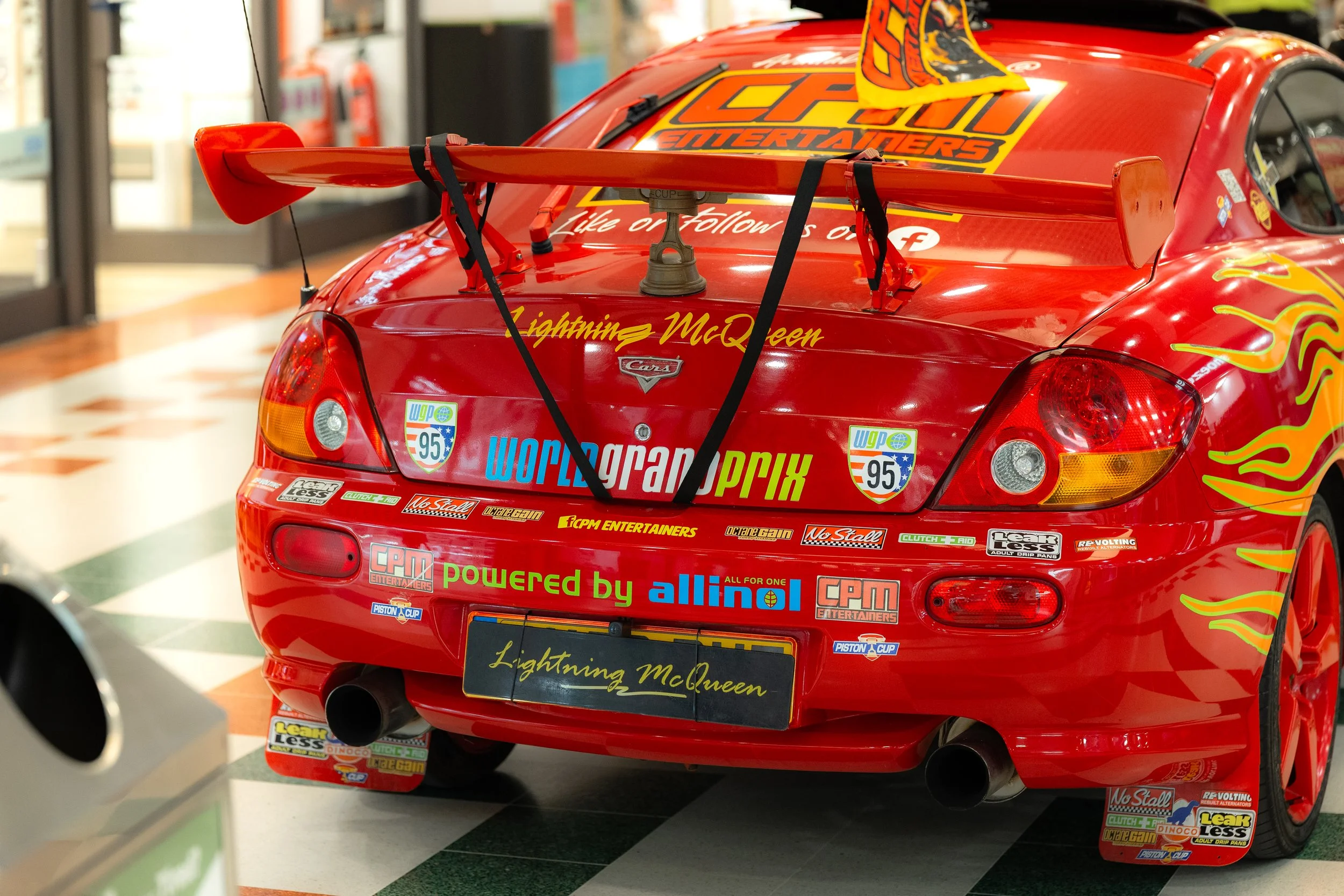 Red race car with various sponsor logos and a large rear wing controlled by black struts, displayed indoors with checkered flooring.