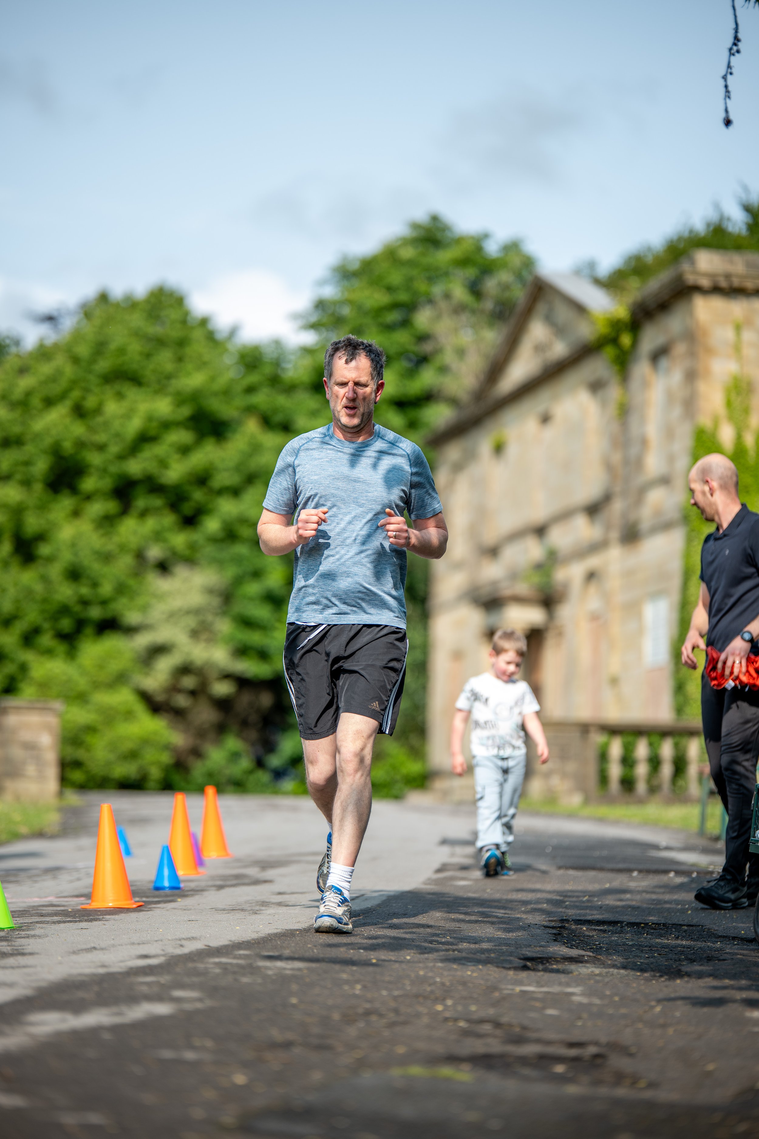 A man running outdoors near traffic cones with a young boy and another man nearby, set against a backdrop of green trees and an old stone building.