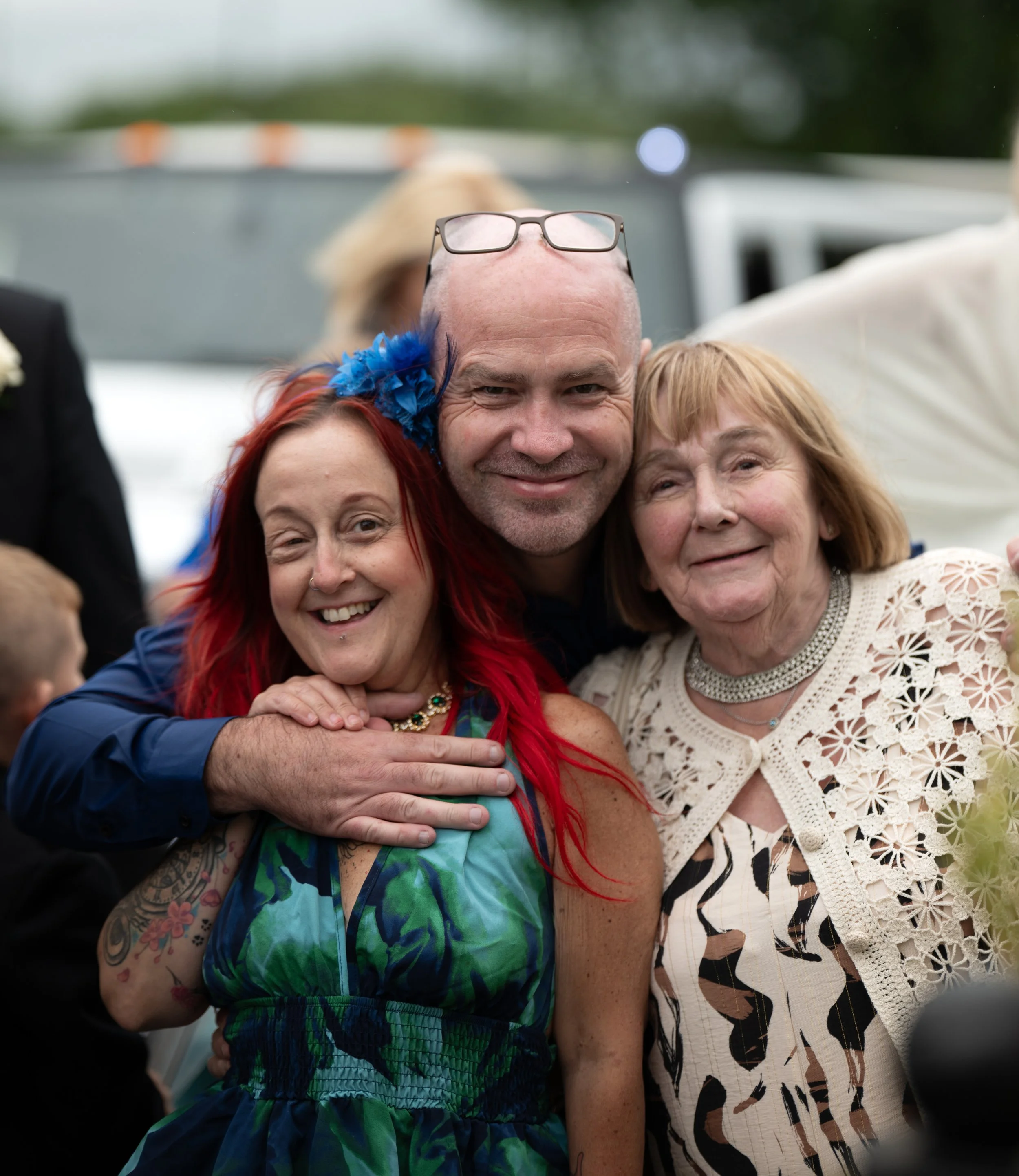 A group of people close together, smiling and embracing each other outdoors. The focus is on a bald man with glasses on his head, surrounded by women with red hair and light brown hair, all appearing joyful during daylight.