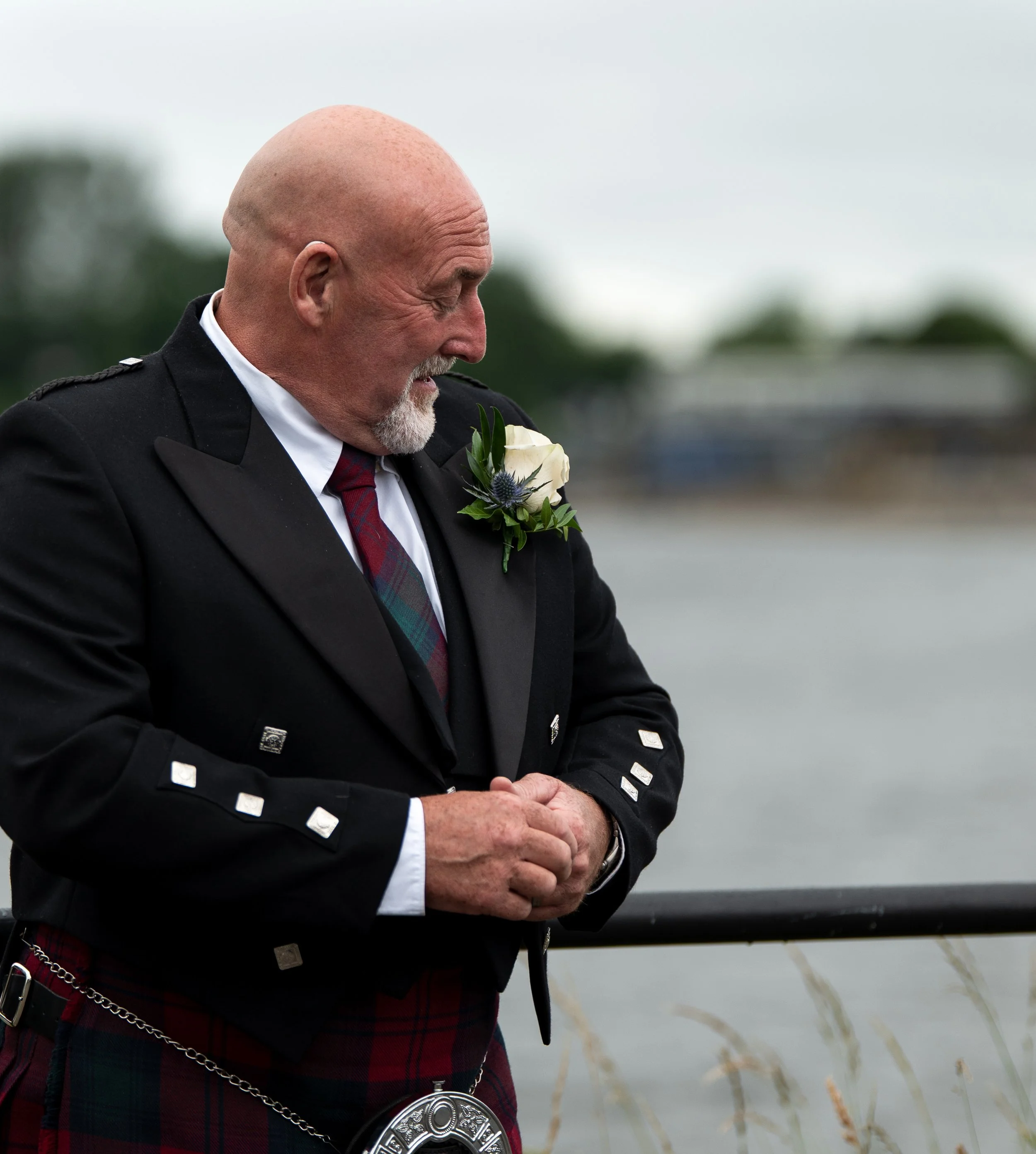 A man in a formal uniform with a badge, white rose, and tartan kilt stands outdoors, with a body of water and reeds in the background.