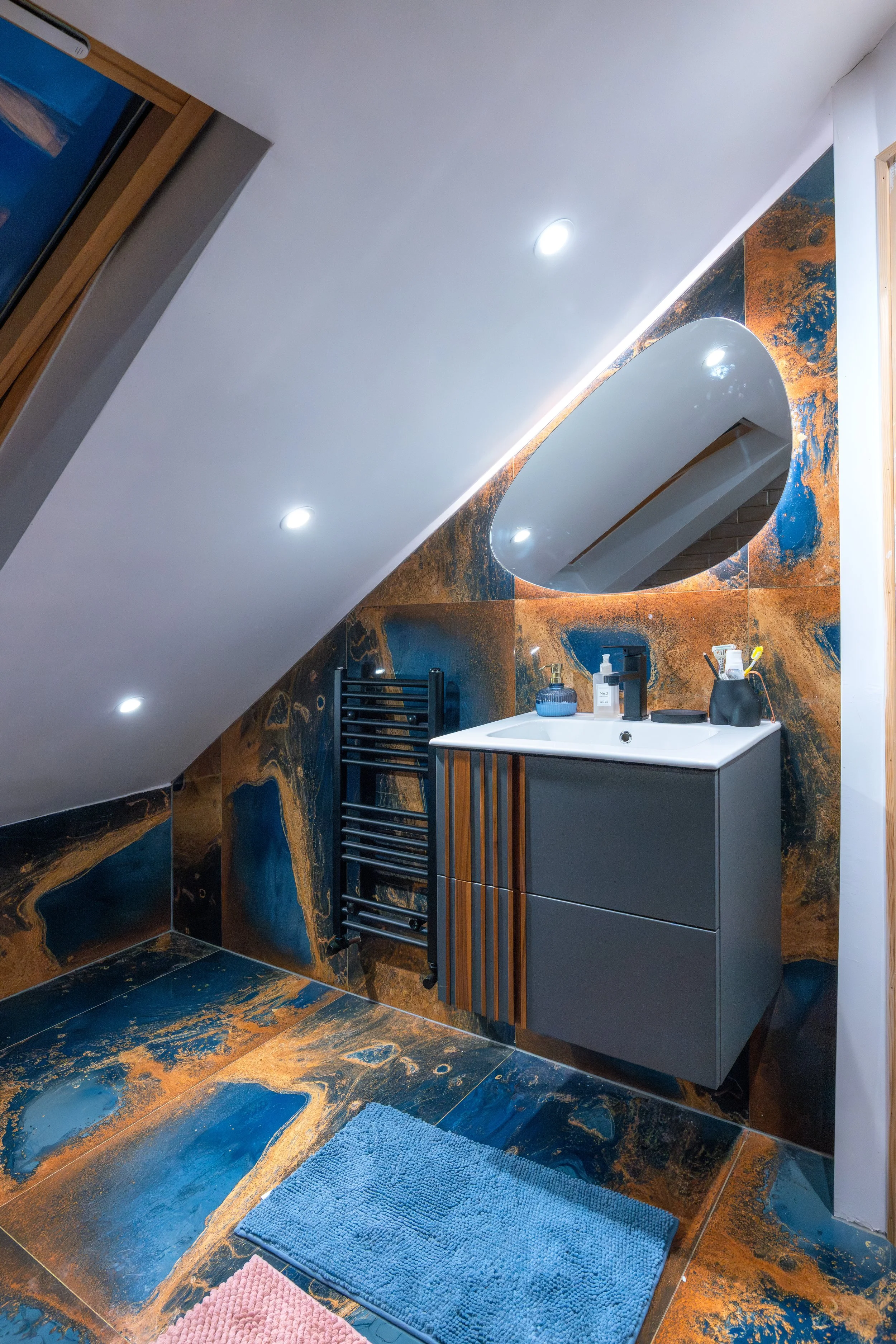 A modern bathroom with dark, marbled tiles on the walls and floor, an oval mirror above the sink, a gray vanity, and a black towel warmer. The ceiling is sloped with recessed lighting, and there are two small rugs on the floor.