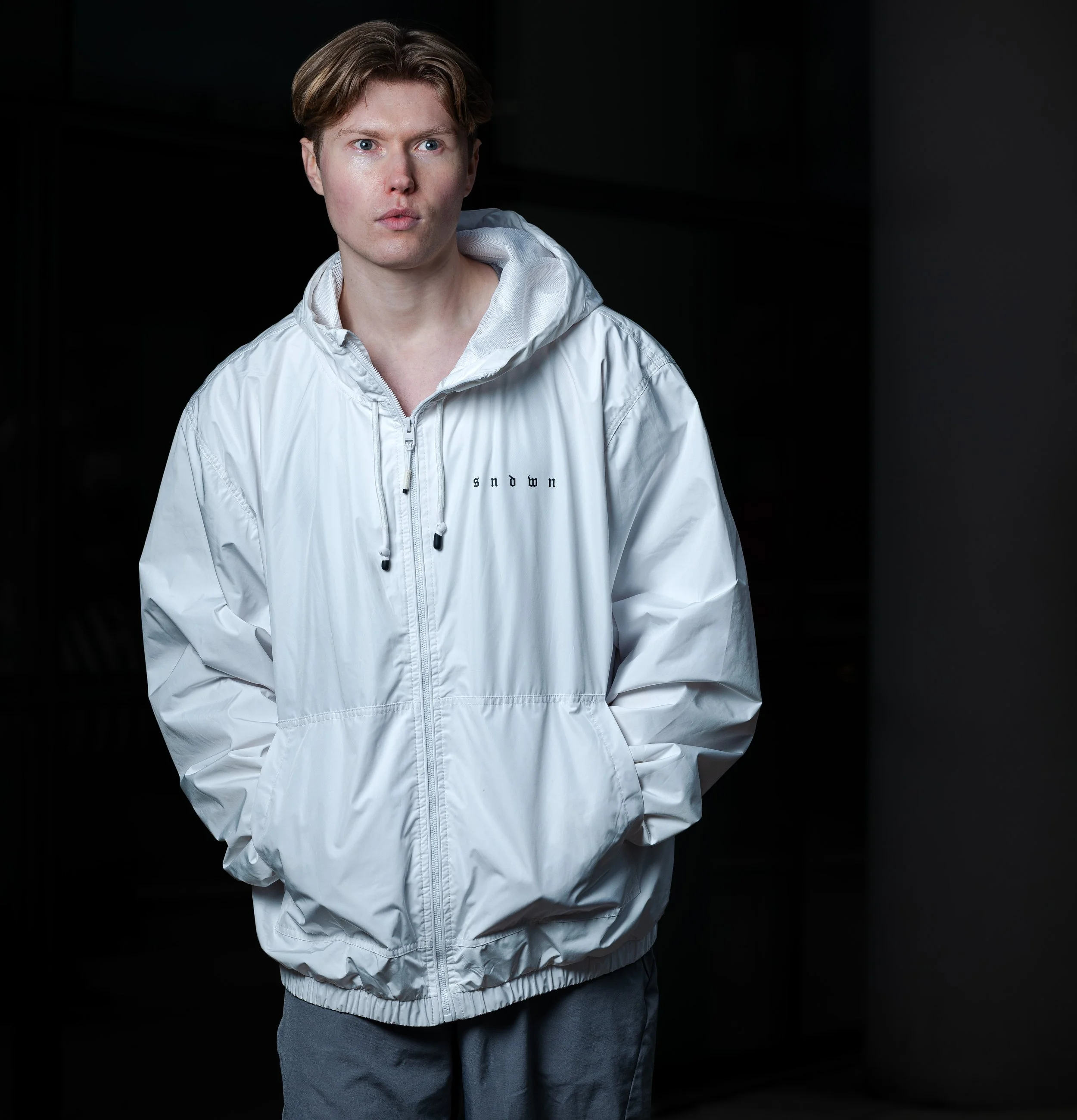 A young man with light brown hair and blue eyes stands in a dark room, wearing a white windbreaker jacket with a hood and a small text logo that says 'S ndwn' on the left chest. He has a serious expression and his hands are in his pockets.