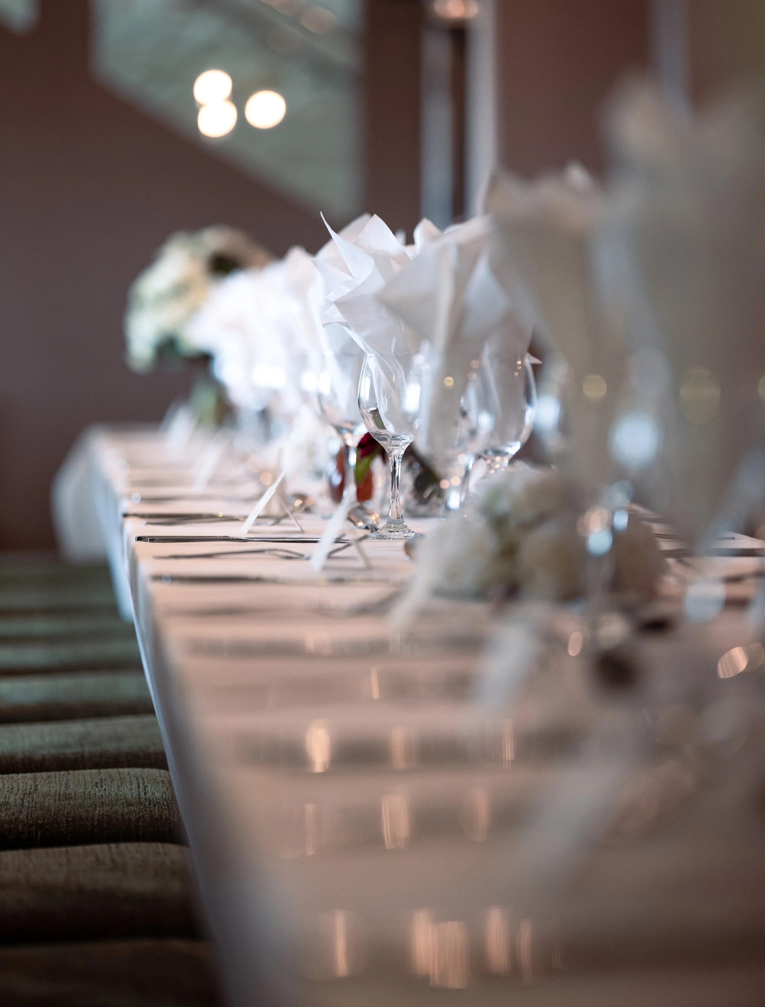 Close-up of a row of empty wine glasses on a table, with white napkins inside each glass, and blurred background of a dimly lit room.