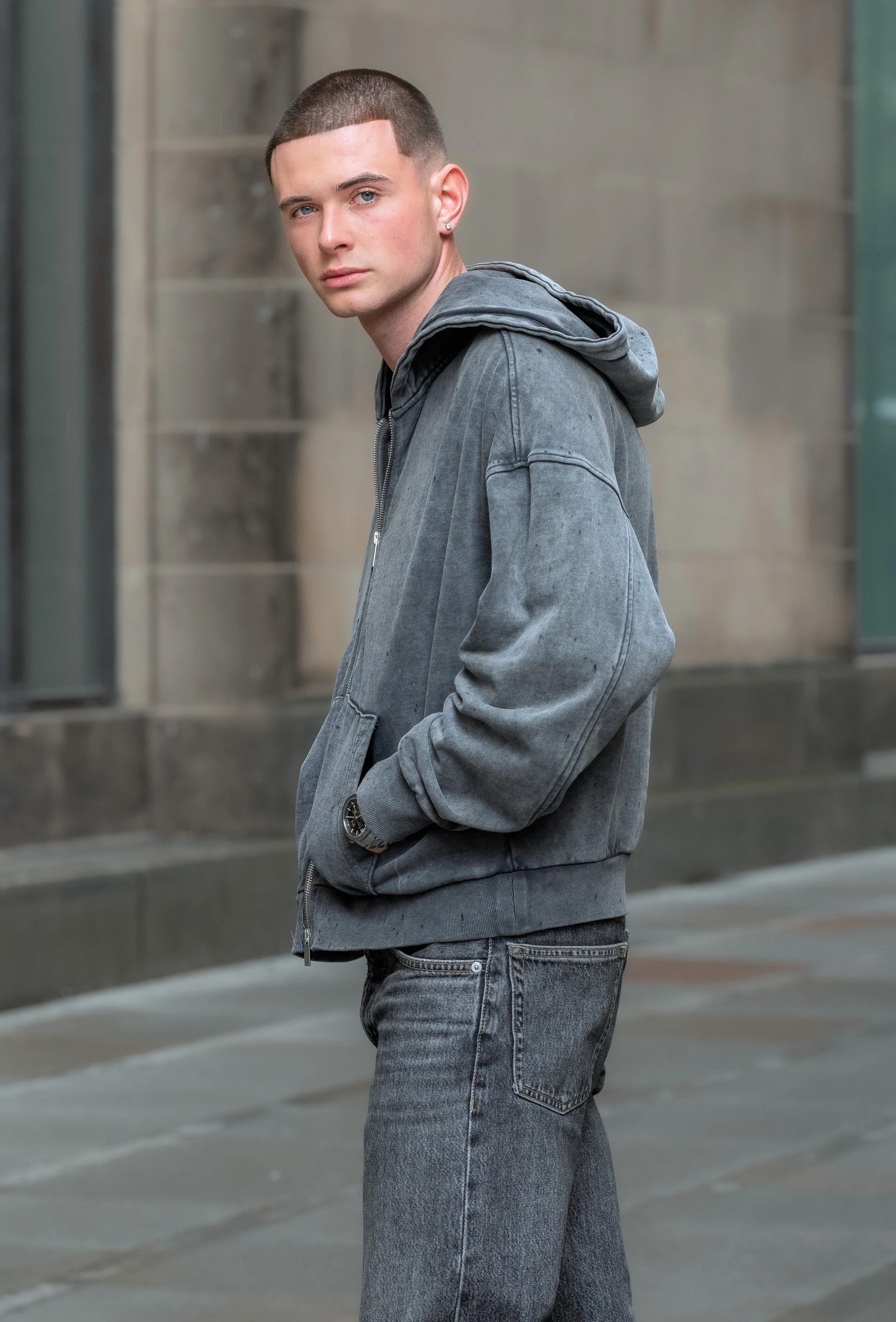 A young man with short hair and blue eyes standing outdoors, wearing a gray hoodie and jeans, with his hands in his pockets, looking at the camera.