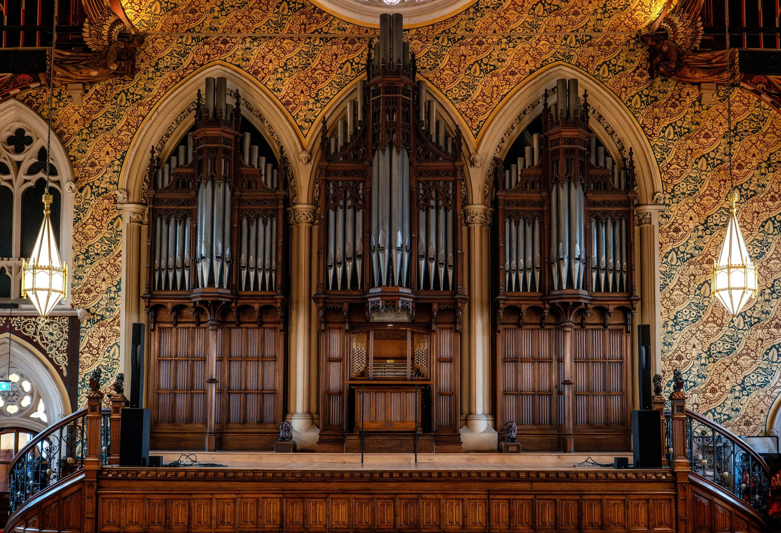 Interior of a church or concert hall with a large pipe organ, ornate woodwork, decorative wallpaper, and hanging lamps.