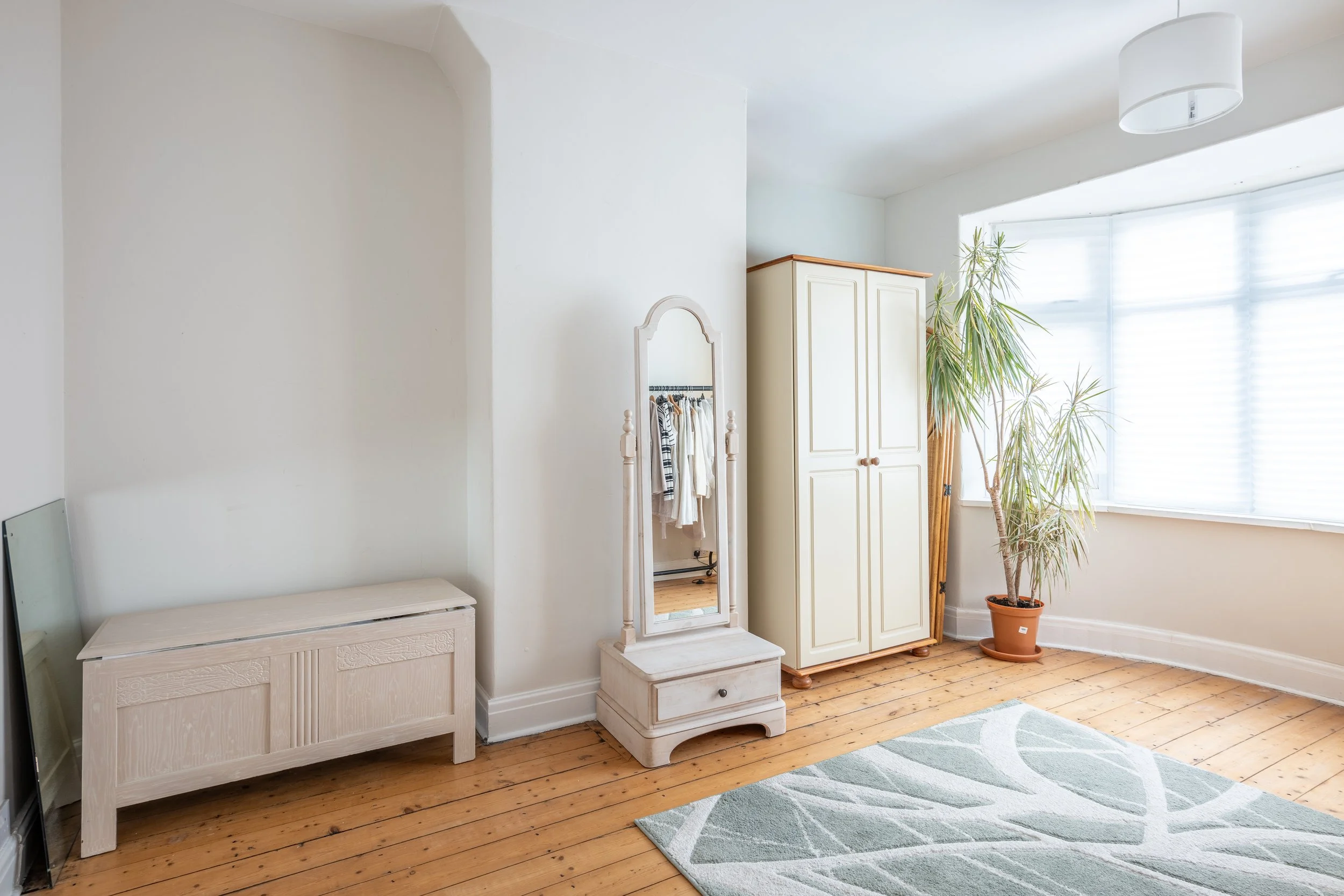 Bright corner of a room with a large window, wooden floor, a potted plant, an oval wardrobe, a full-body mirror, a light-colored storage bench, and a green and white patterned rug.