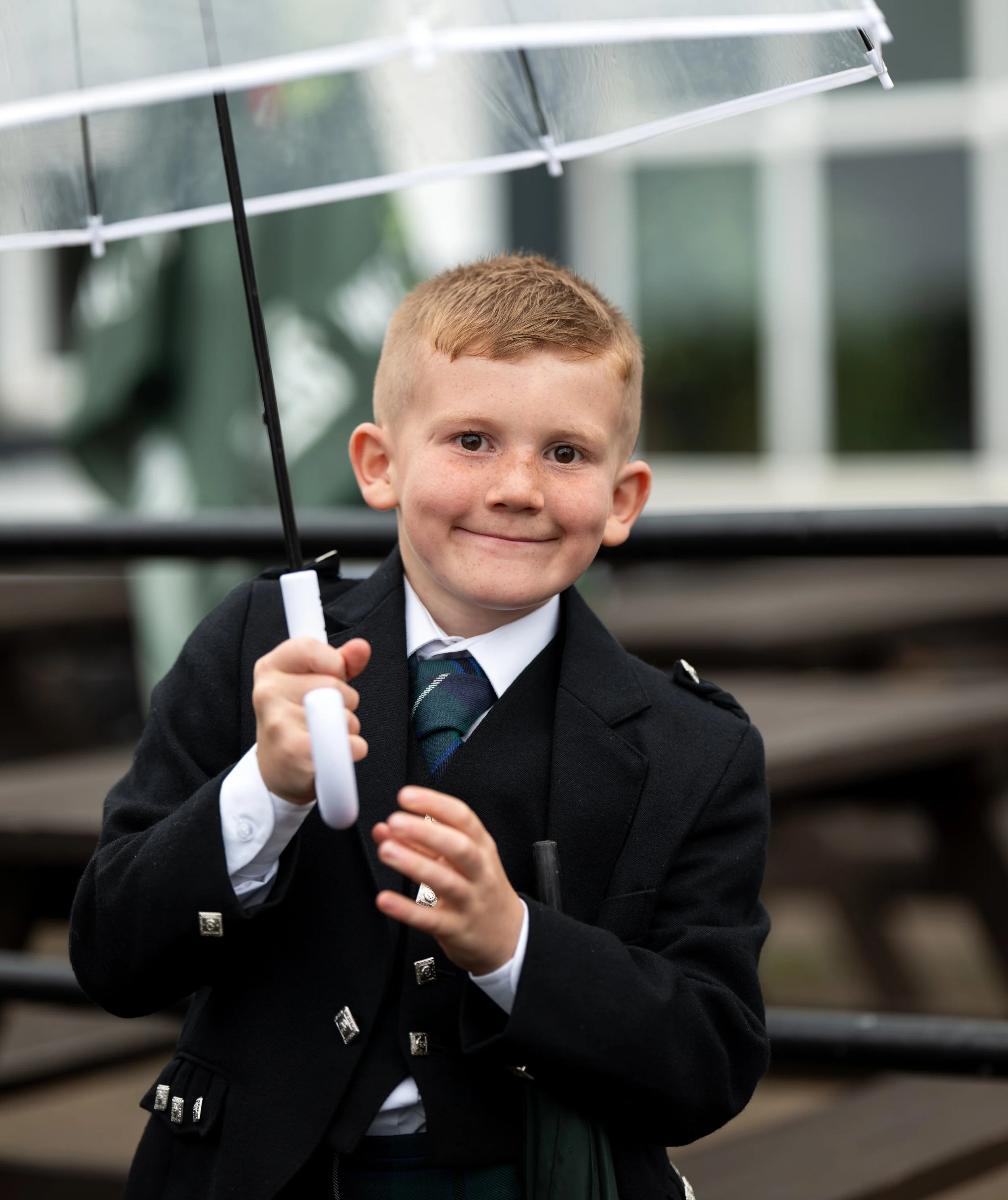 A young boy with red hair and freckles smiling and holding a white marker, standing outdoors in front of metal bleachers, wearing a black blazer, white shirt, and a tartan tie.