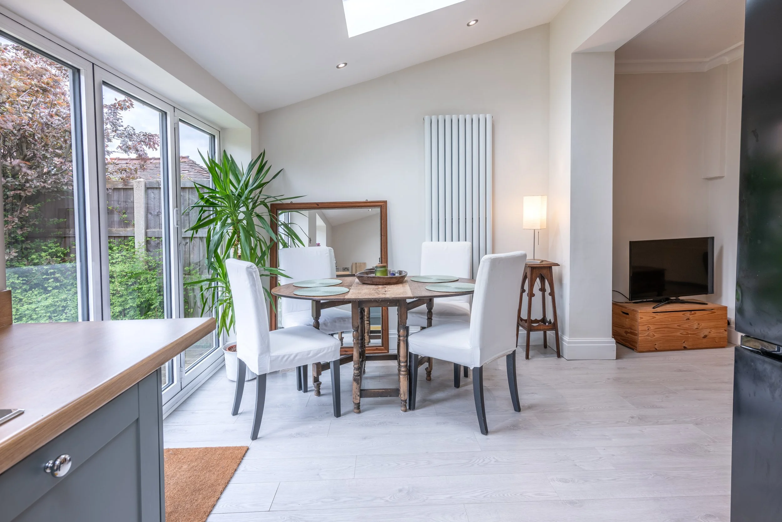 Photo of a dining area with a round wooden table and five white chairs, adjacent to large sliding glass doors leading to an outdoor garden. The room is bright with natural light and has light-colored wooden flooring, a ceiling skylight, a tall radiat