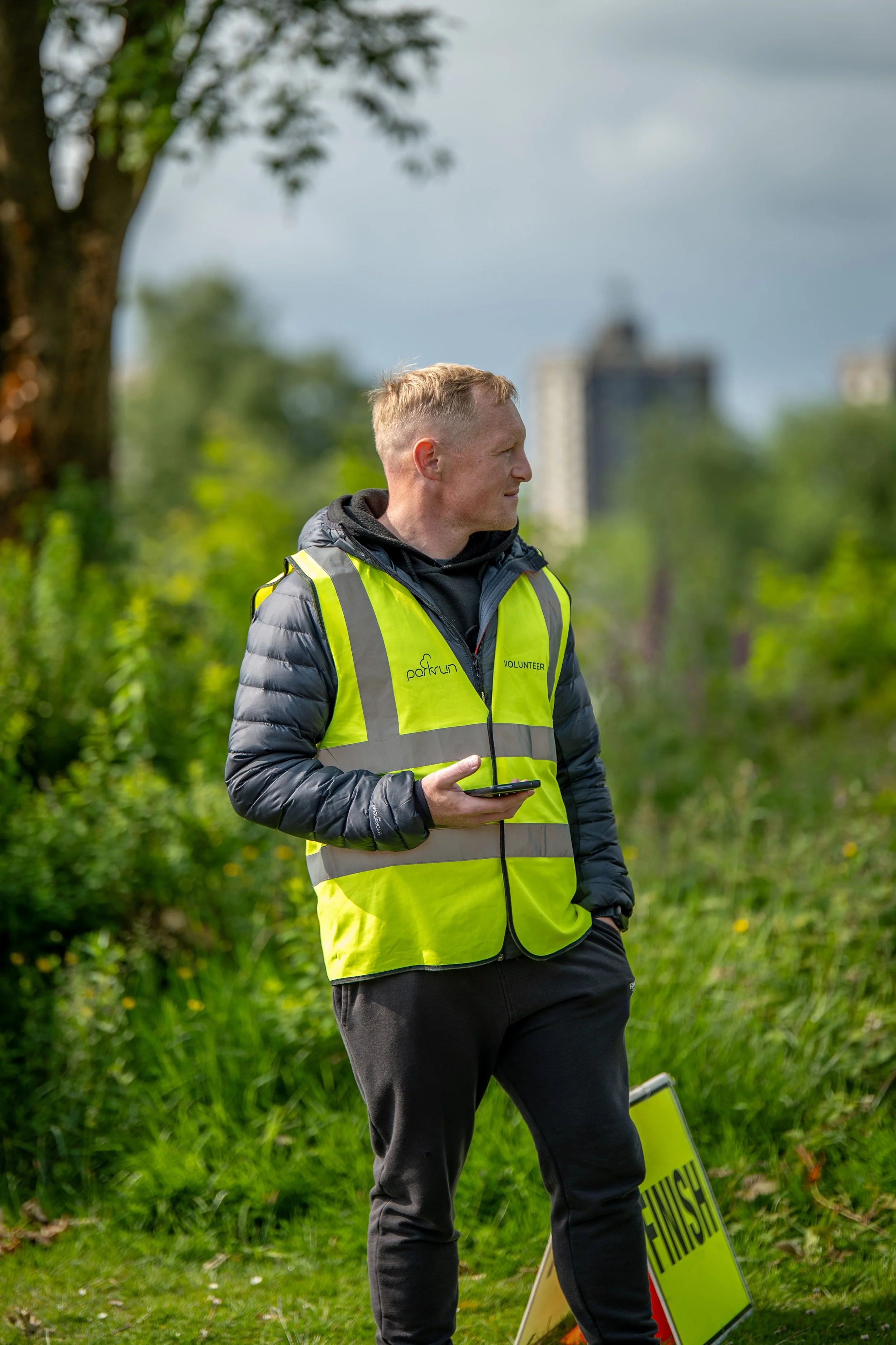 A man wearing a yellow safety vest with the words 'Parkrun' and 'Volunteer' stands outdoors on a grassy area, holding a smartphone. There is a finish line sign nearby, trees, and high-rise buildings in the background.