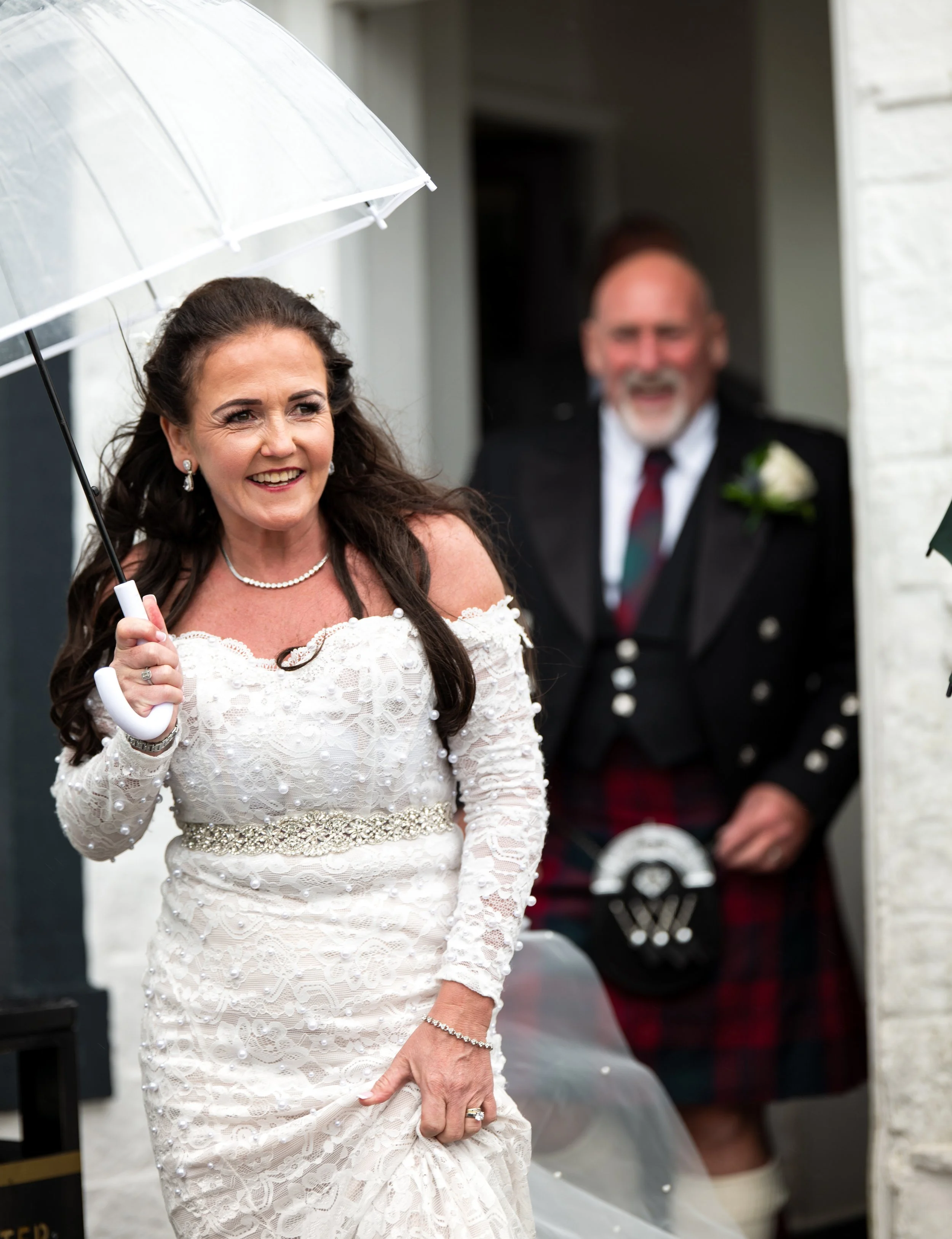 A smiling woman in a white lace wedding dress holding a clear umbrella during a wedding ceremony, with a man in traditional Scottish attire in the background.
