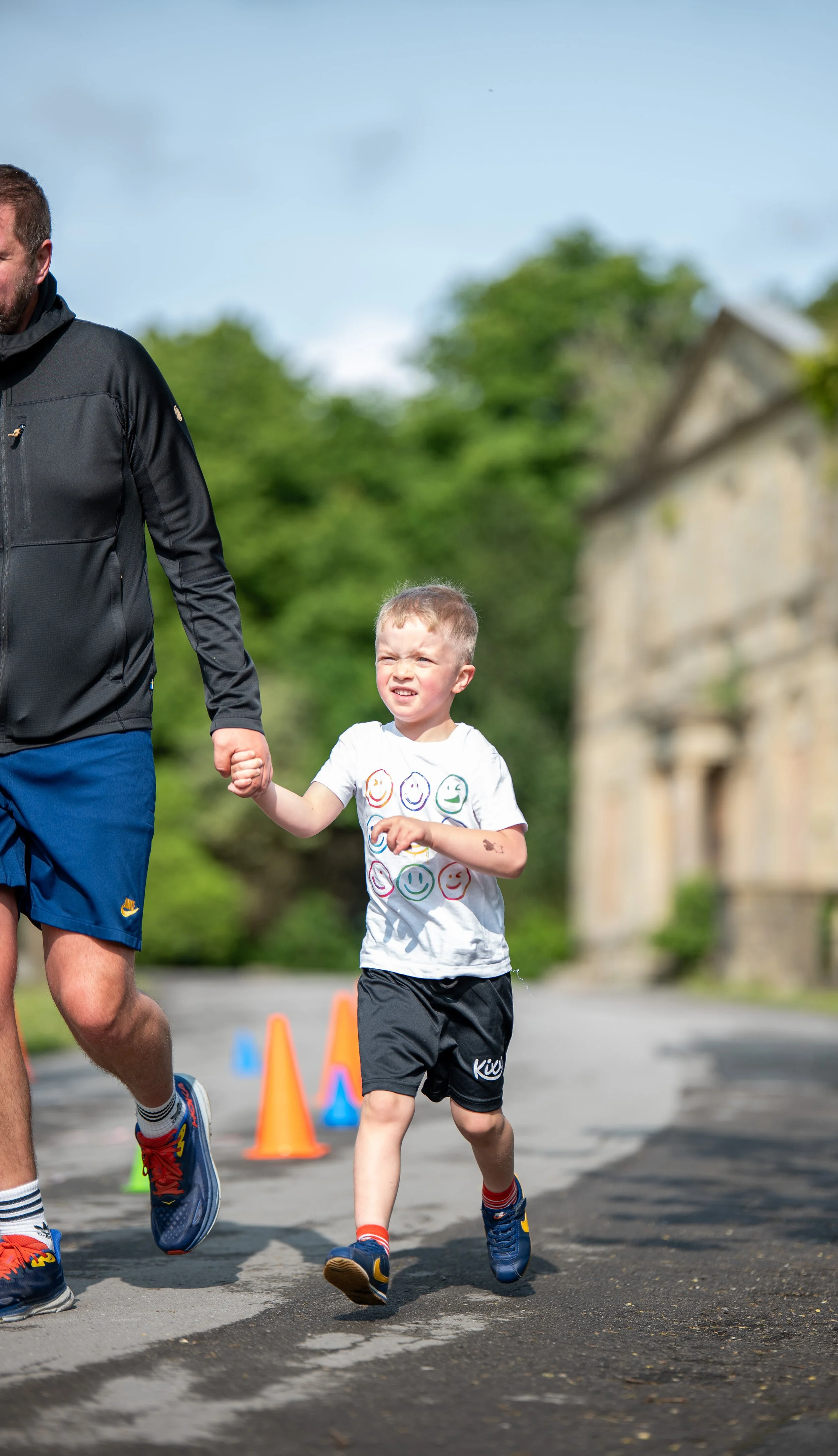 A young boy running with an adult, holding hands, on a road marked with orange traffic cones, with trees and an old building in the background.