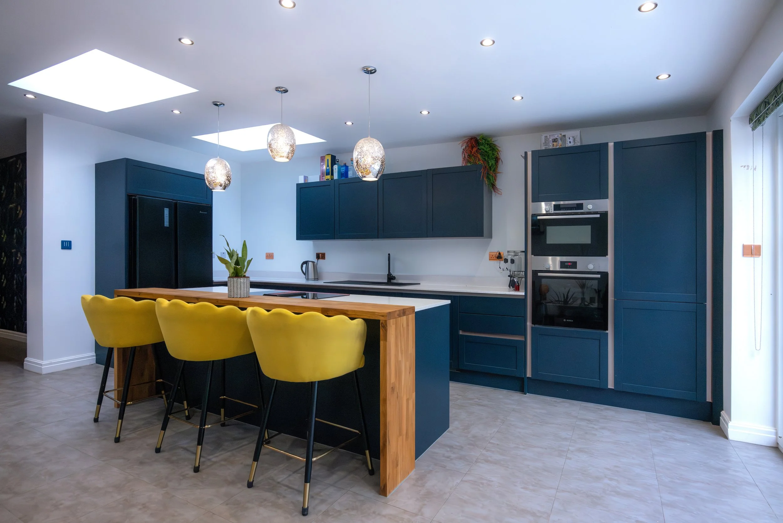 Modern kitchen with navy blue cabinets, white countertops, yellow bar stools, and a wooden kitchen island. Recessed lighting and three pendant lights hanging over the island, with skylights on the ceiling.