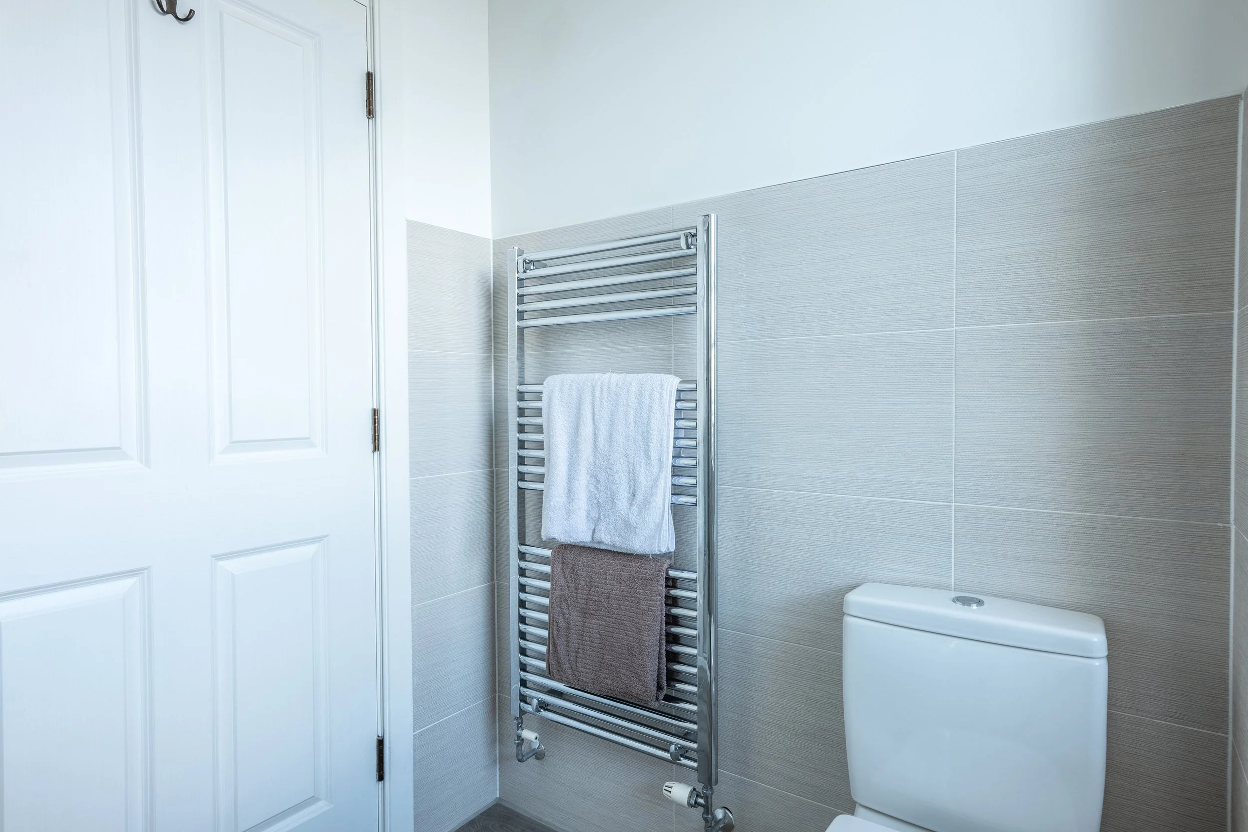 A modern bathroom with beige tiled walls, a white toilet, and a chrome towel radiator with three towels hanging from it. There is a closed white door on the left side.