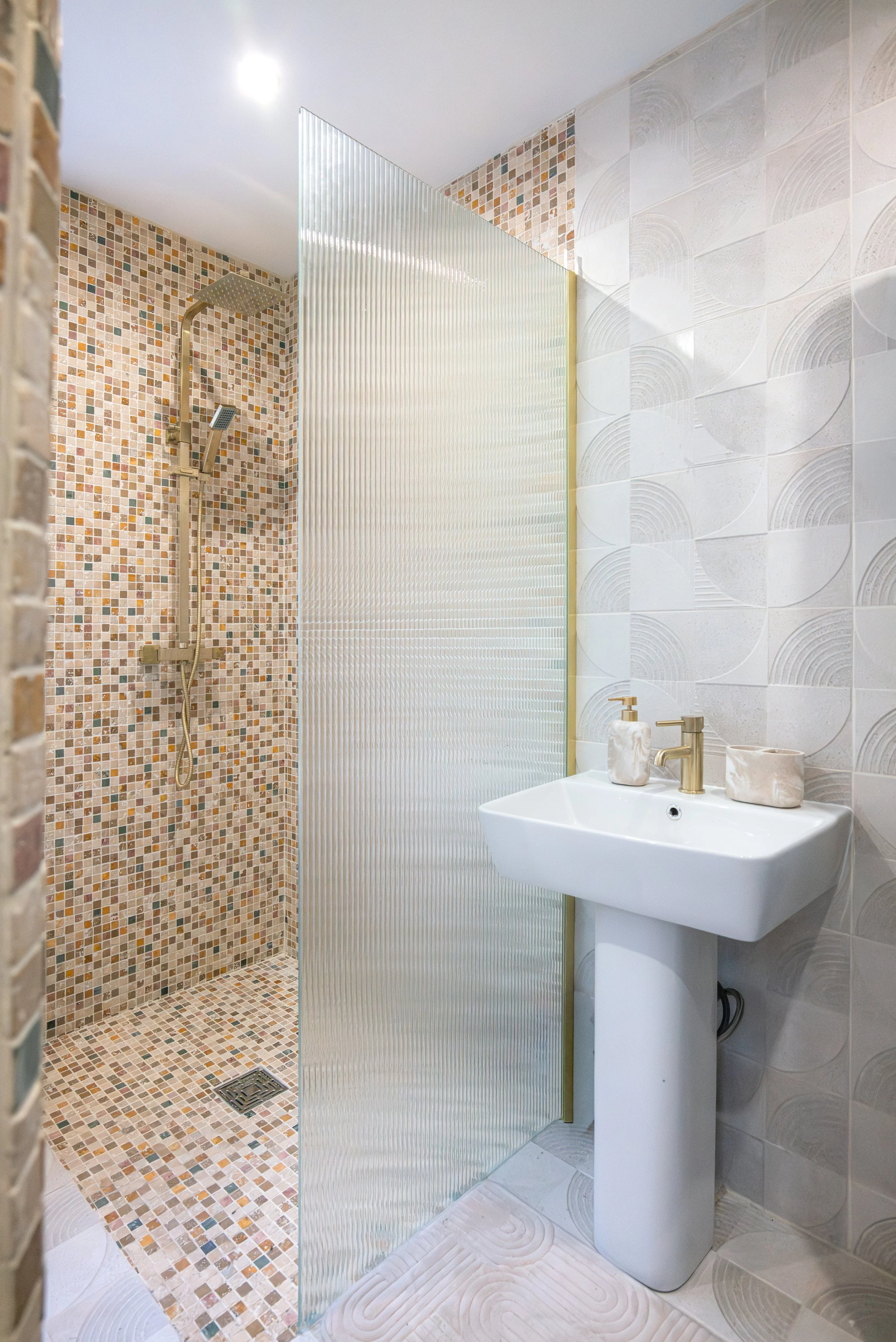 Bathroom with a walk-in shower featuring small mosaic tiles, a frosted glass partition, a white pedestal sink, and decorative wall tiles.