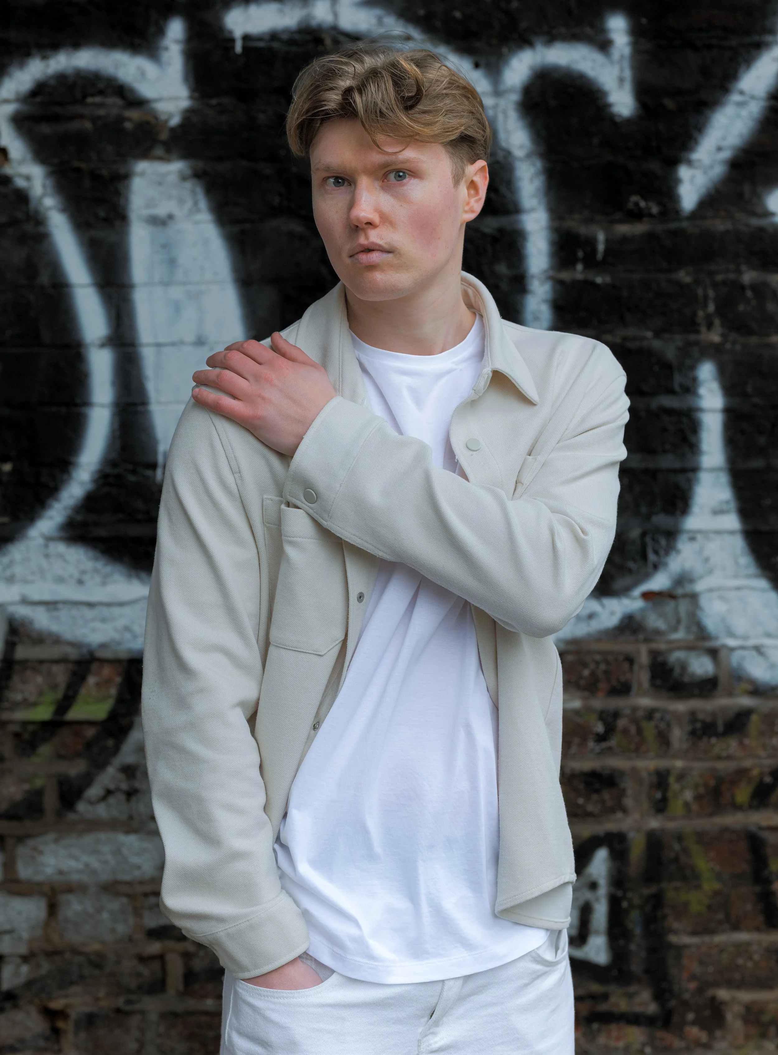 A young man with light brown hair and blue eyes standing in front of a brick wall with graffiti. He is wearing a white T-shirt and a light beige jacket, with one hand resting on his shoulder and the other in his pocket.