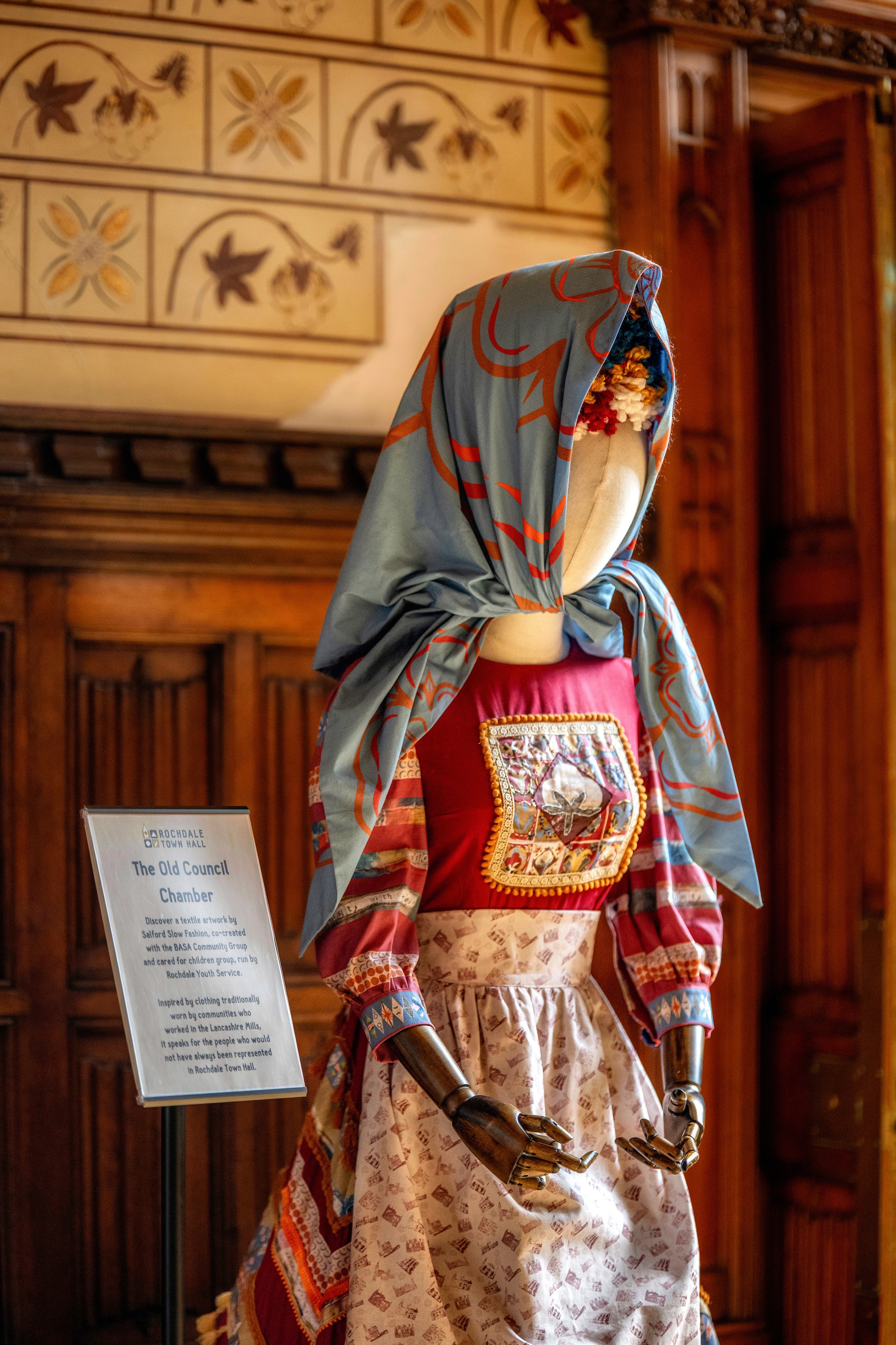 A mannequin dressed in traditional clothing, including a colorful dress with a decorated patch on the chest, a wide pink waistband, and a blue headscarf with red and gray patterns. The mannequin is displayed indoors against a wooden wall with decorat