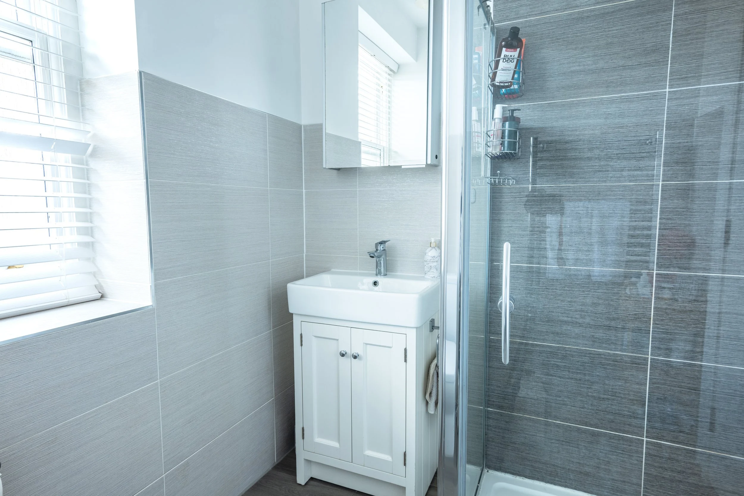 Small bathroom with a white sink cabinet, a mirror, a soap dispenser, and a glass-enclosed shower with shelves holding shampoo and body wash bottles. Window with blinds on the left
