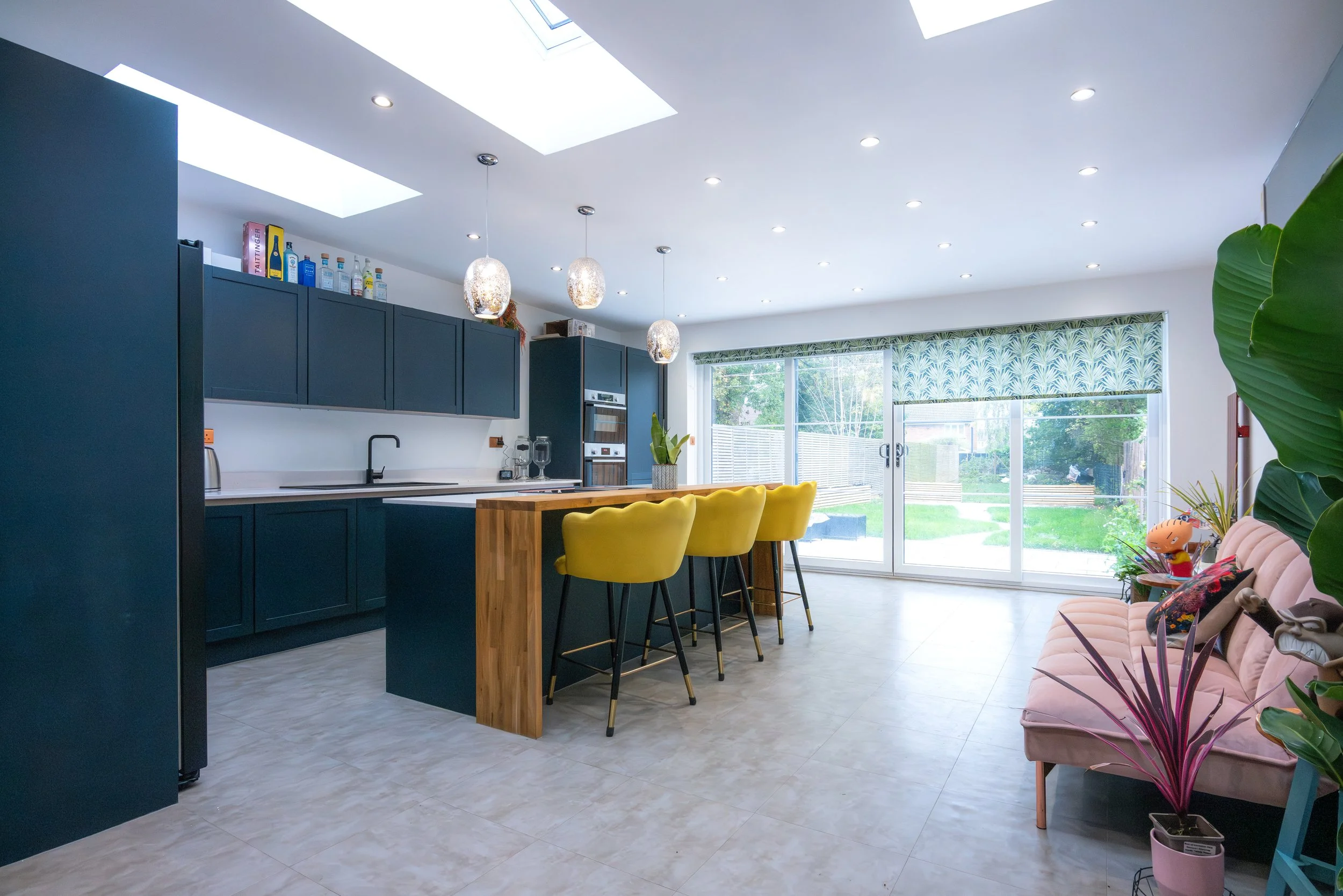 Modern kitchen and living area with navy blue cabinets, a wooden island with yellow barstools, and large sliding glass doors showing a backyard garden.
