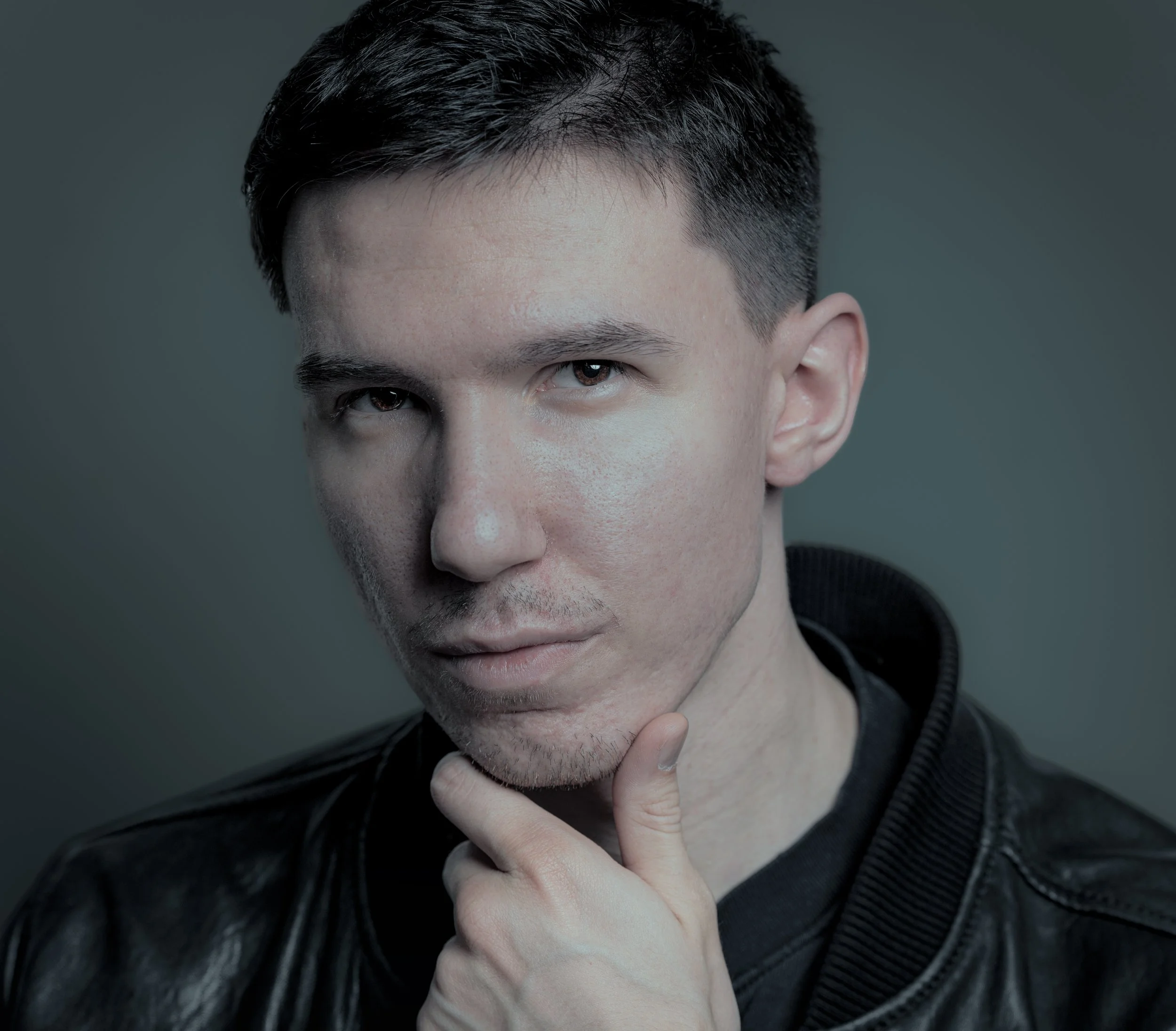 A young man with short dark hair and light skin, wearing a black leather jacket, posing with his chin resting on his hand against a plain gradient background.