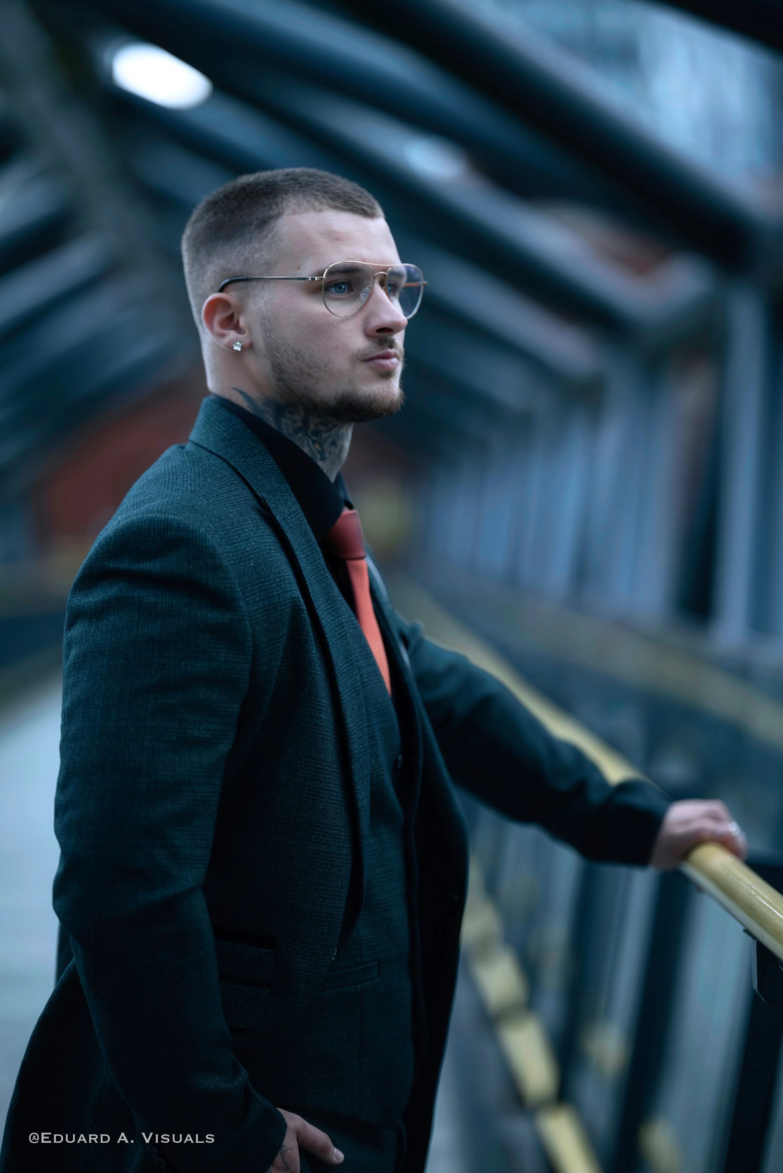 A young man with short hair and glasses standing on a bridge or walkway, looking off to the side, wearing a dark blazer, black shirt, and red tie, with a tattoo visible on his neck, holding onto a yellow handrail.