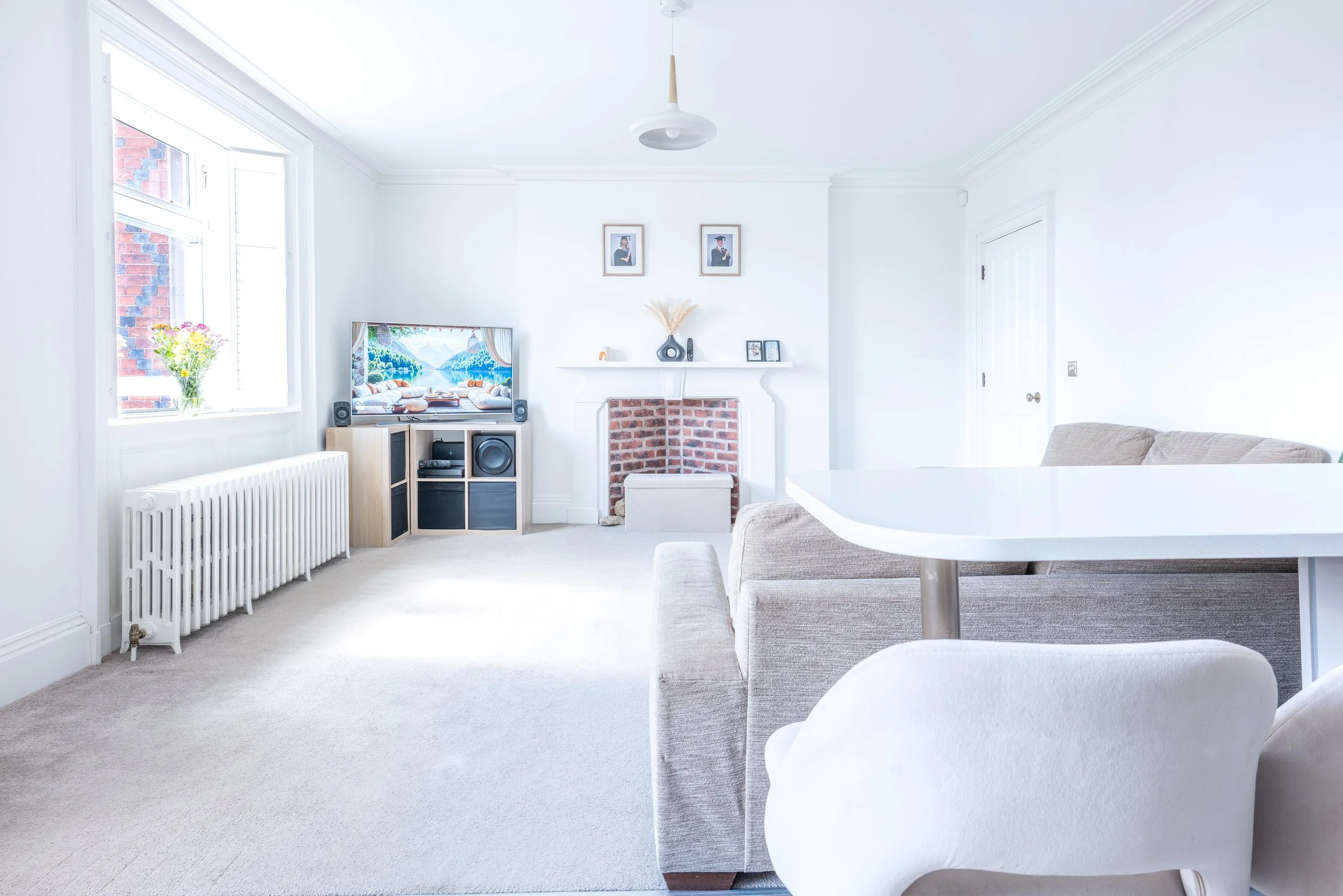 Bright, white living room featuring a TV with landscape scenery, framed photos, a small fireplace, a vase with dried flowers, a vinyl speaker system, a grey sofa, and a white round dining table.