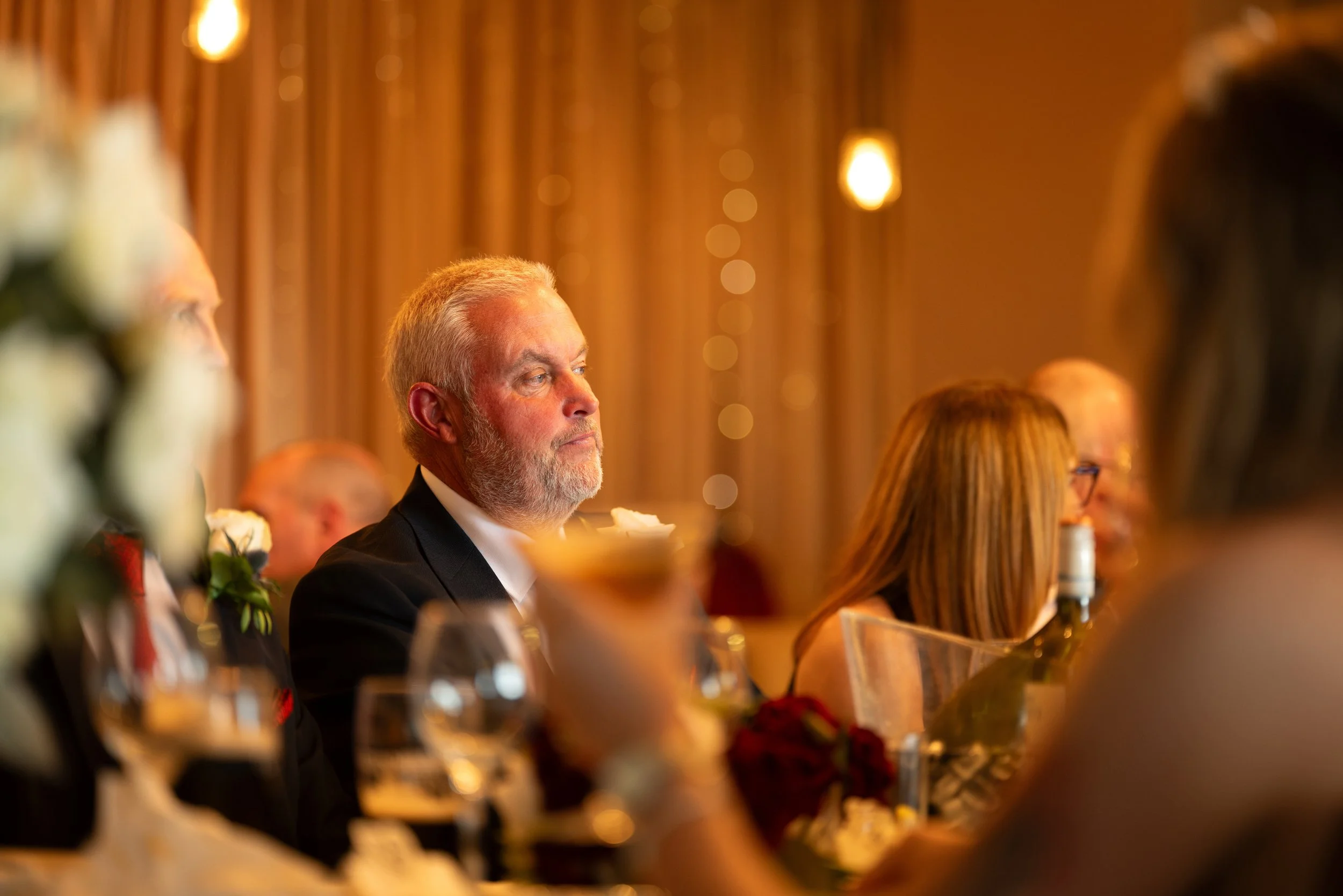 A group of people seated at a formal event, with a man with gray hair and a beard in focus, wearing a suit, with blurred background and warm lighting.