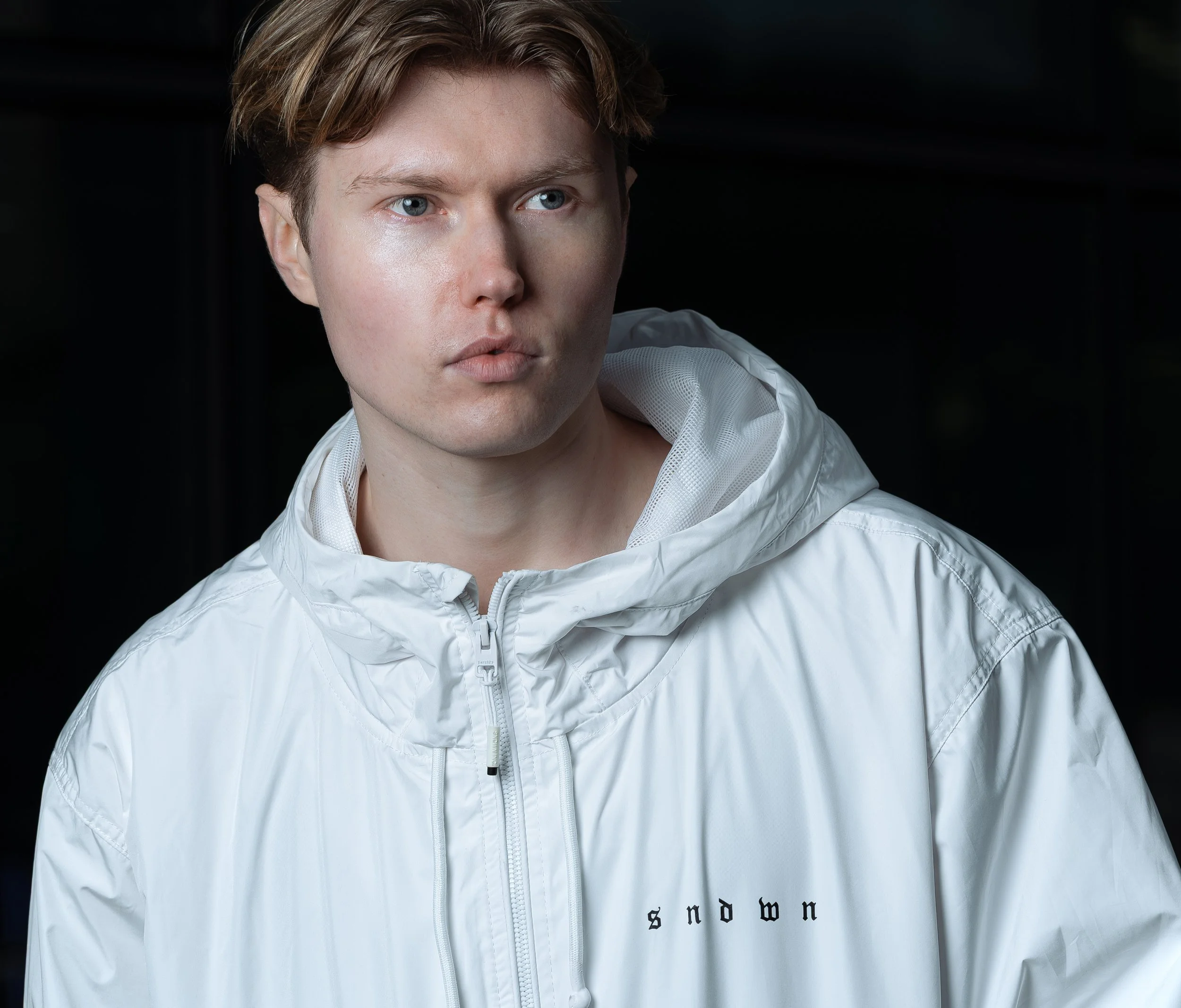 Young man with light skin and reddish-brown hair wearing a white hooded jacket with the word 'snow' on it, looking to the left against a dark background.