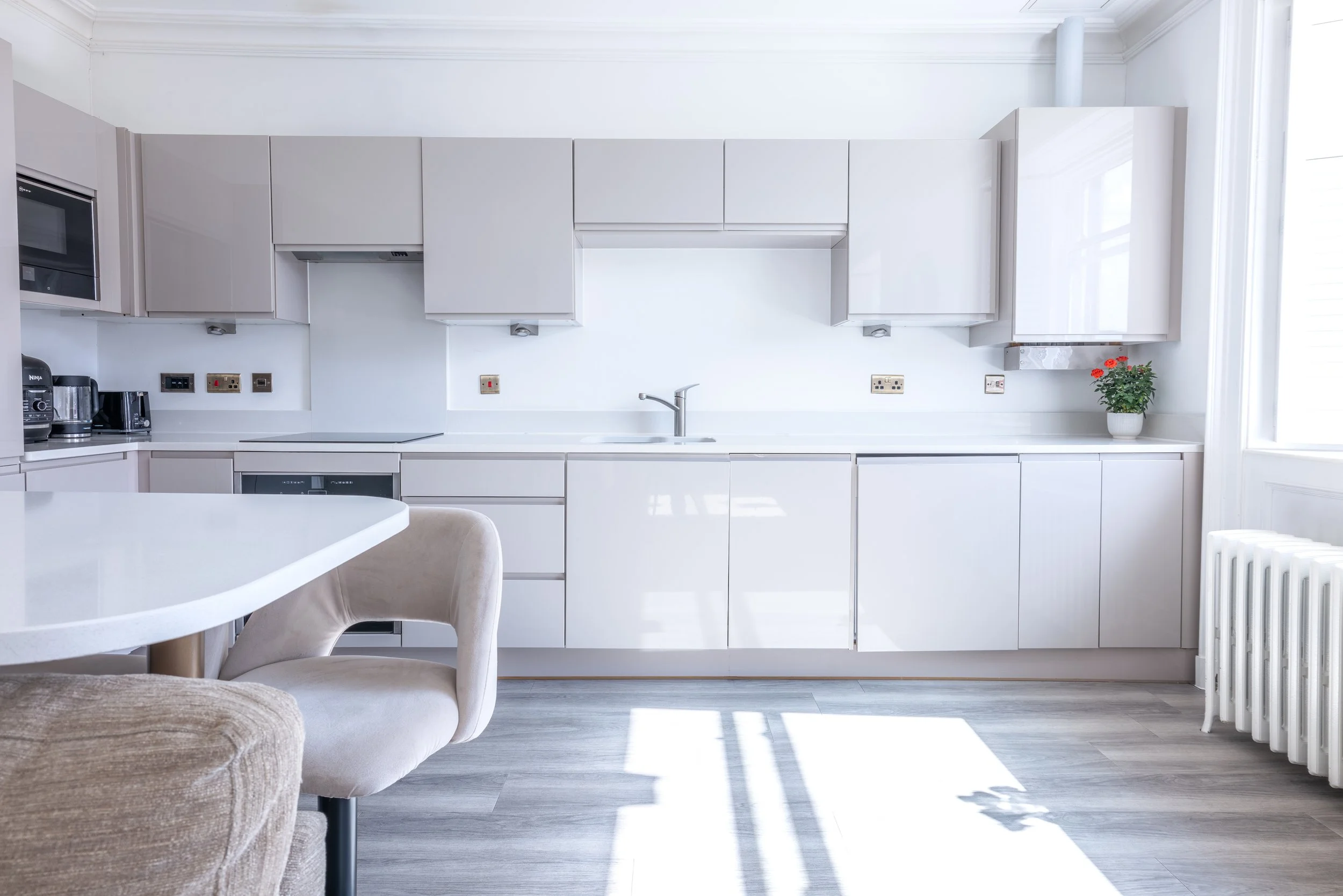 A modern, minimalist kitchen with white cabinets, a white countertop, a stainless steel sink with a faucet, and a small potted red flowering plant on the counter. There is sunlight streaming in through a window on the right, creating shadows on the f