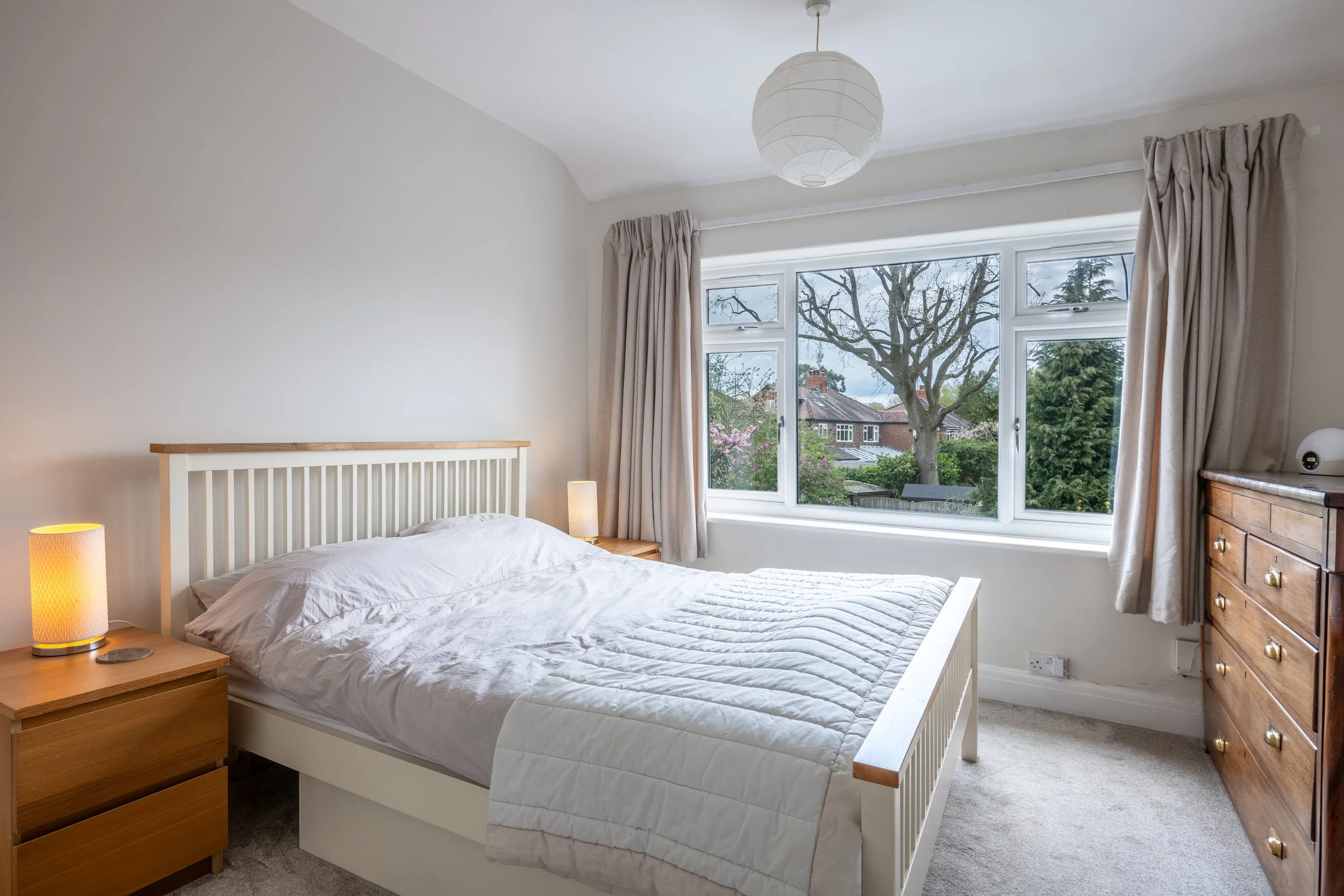 A bedroom with a white bed, a wooden nightstand with a lit lamp, a large window with beige curtains showing a tree and neighboring houses, and a wooden dresser with a white speaker on top.