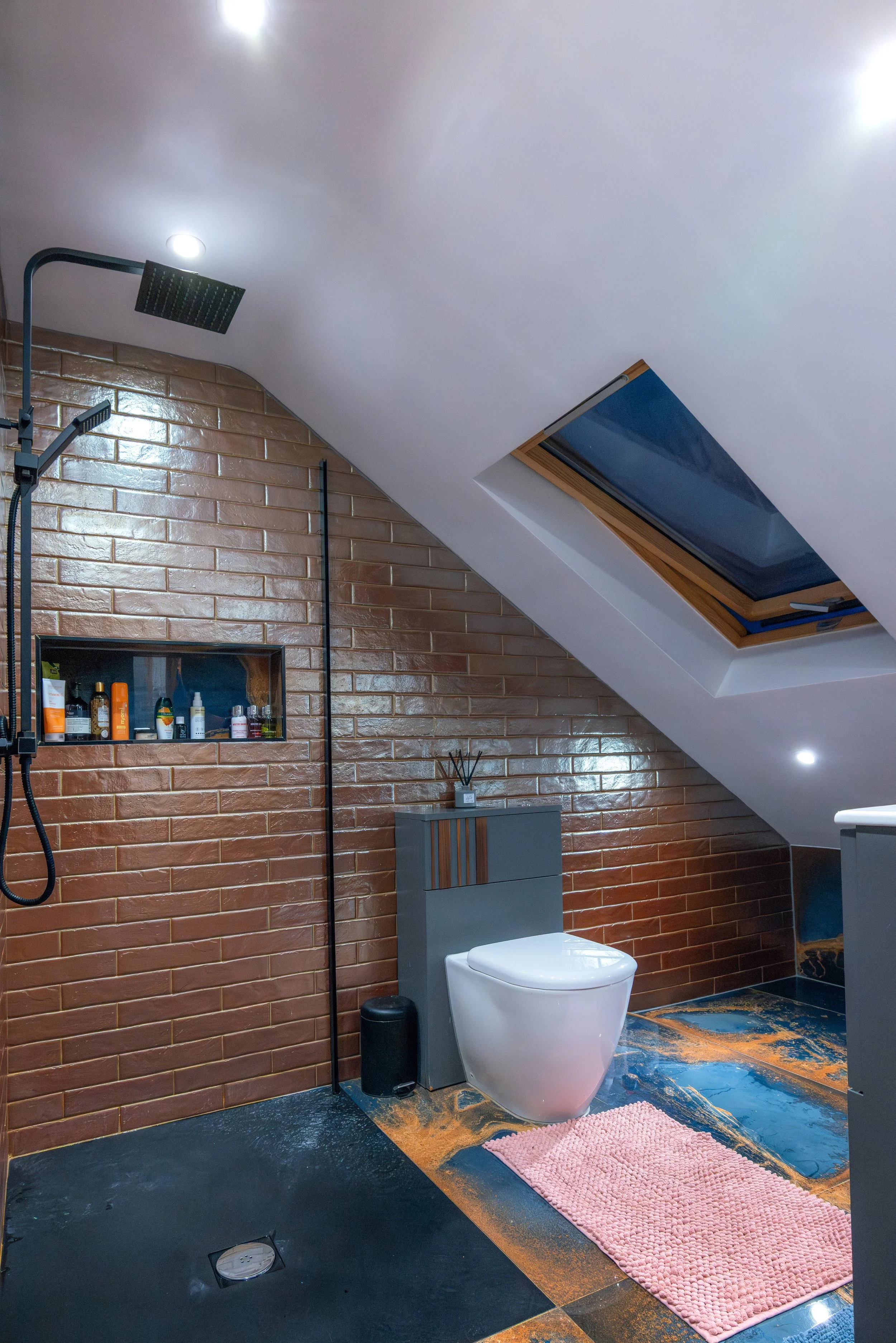 Modern bathroom with sloped white ceiling, skylight, brick accent wall, shower area with rainfall showerhead, toilet, pink bath mat, and various toiletries on a built-in shelf.