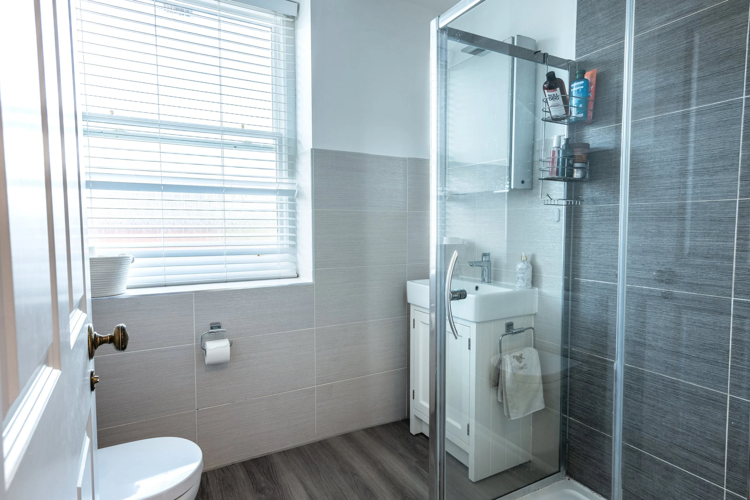 A bathroom with a window with blinds, a toilet, a small sink, a mirror, and a shower enclosed by glass with various toiletries on shelves.