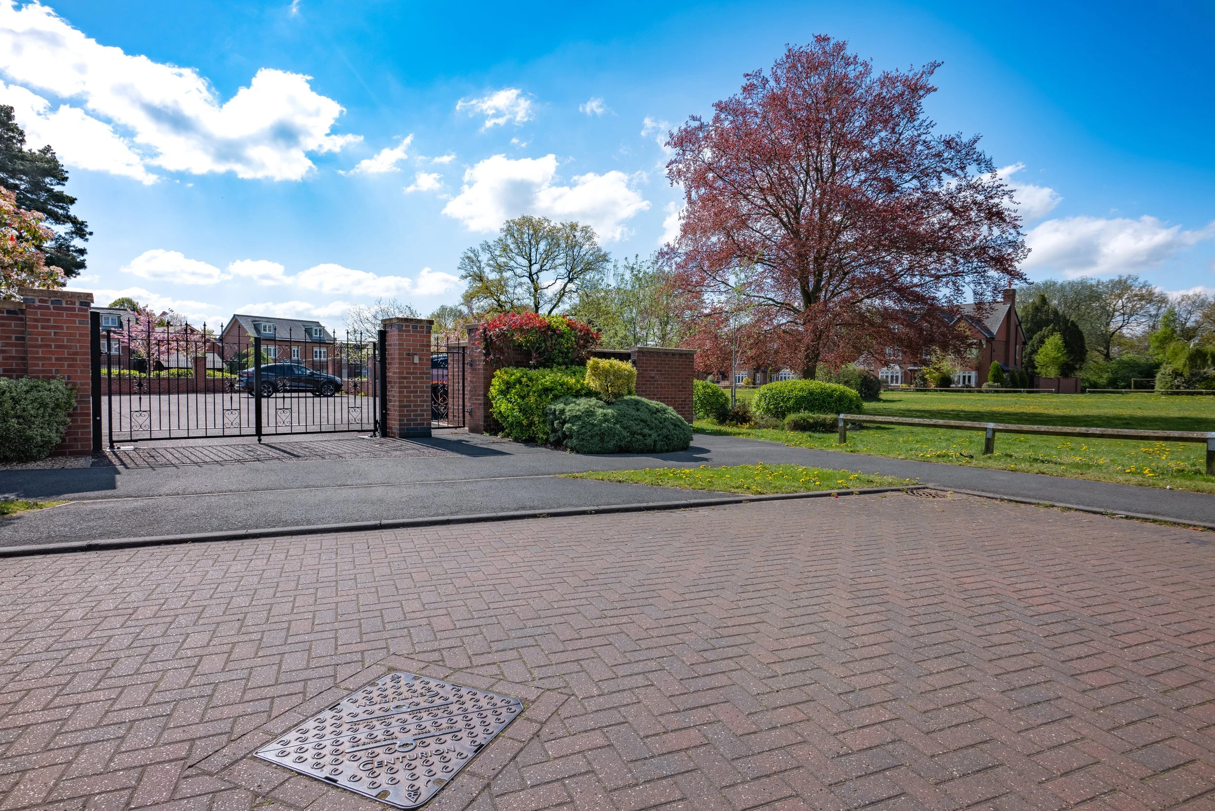 Street view showing a sidewalk, a gated entrance to a residential area with brick columns, trees with spring foliage, and houses under a partly cloudy blue sky.