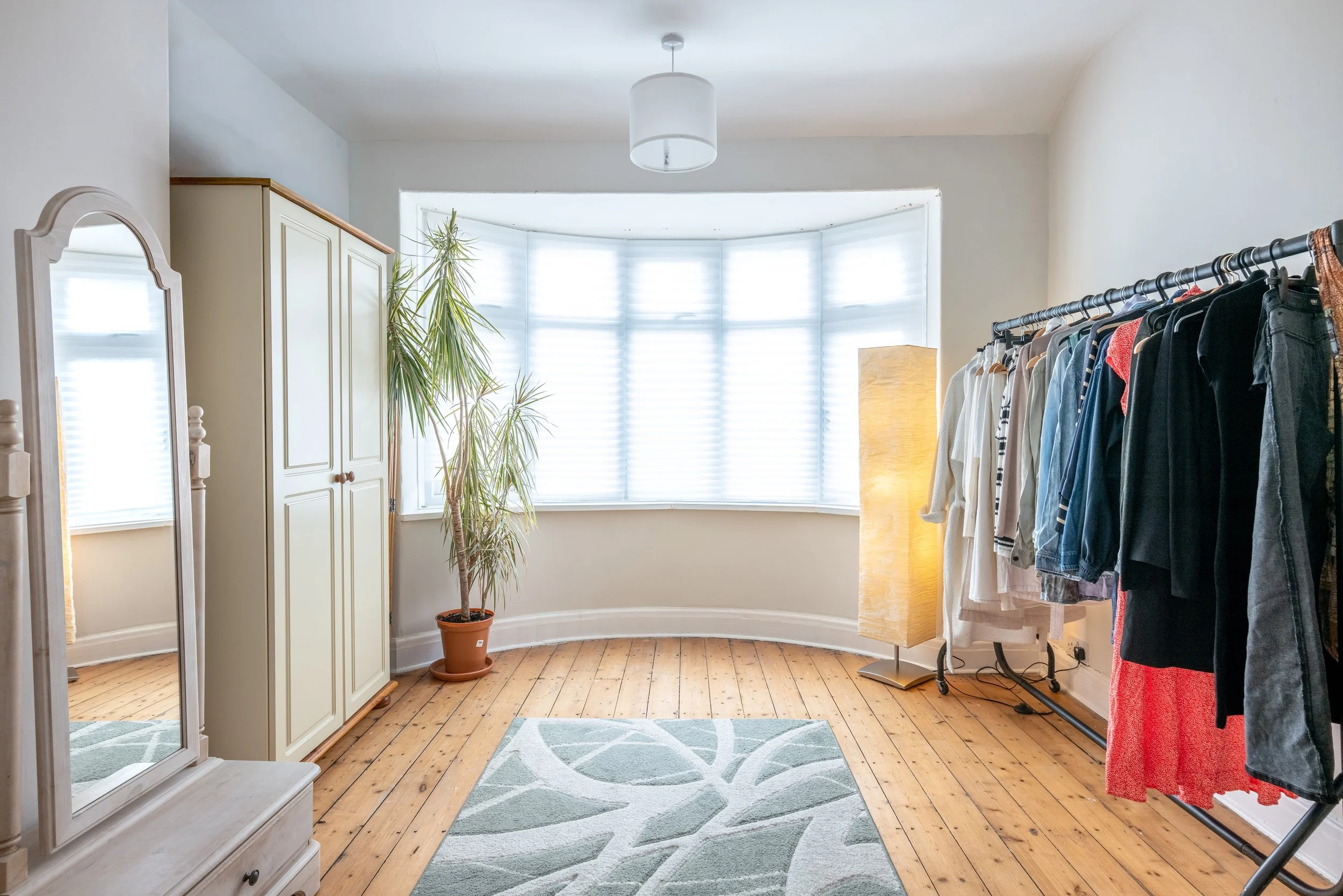 Bright room with large bay window with blinds, wooden floor, a clothing rack with various clothes, a tall potted plant, a lamp, a mirror, and a patterned rug.