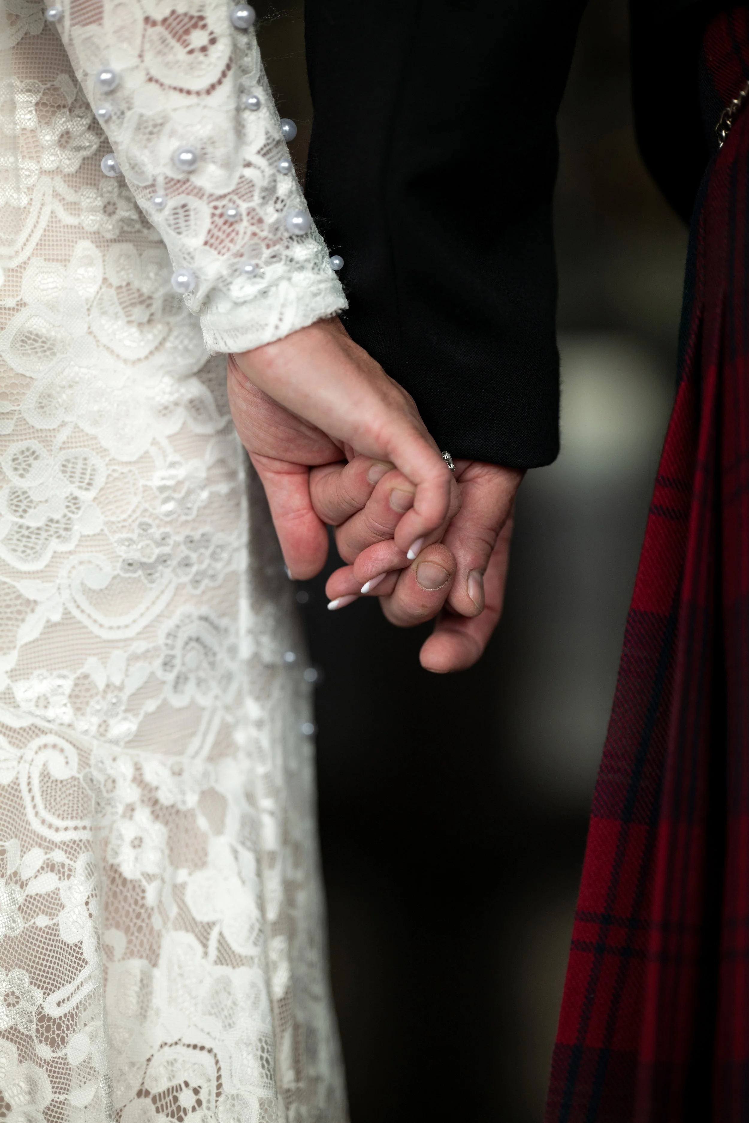 Close-up of a couple holding hands, dressed in wedding attire, with the bride wearing a lace dress with pearl embellishments and the groom wearing a dark suit.