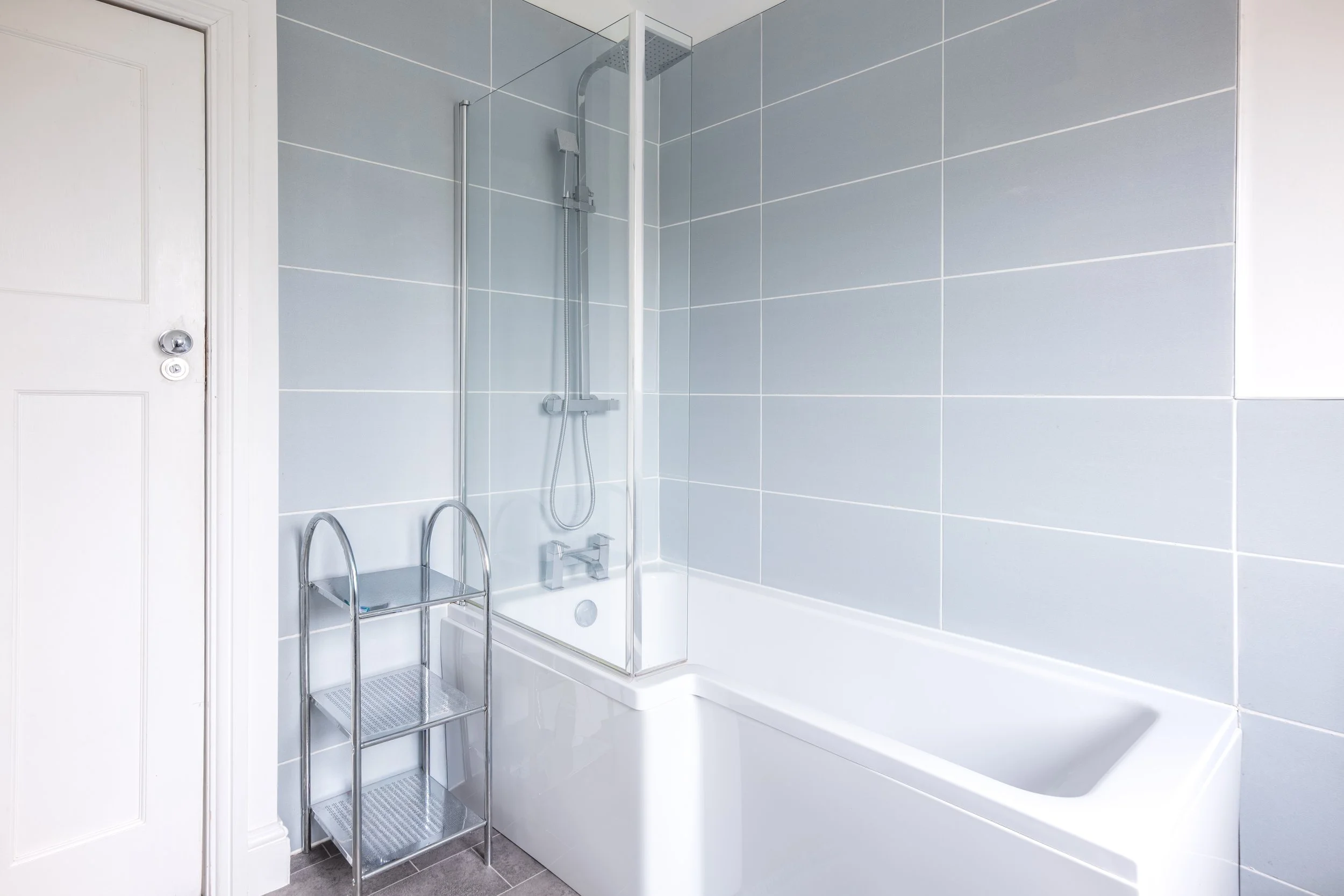 Bathroom with light blue tiled walls, white bathtub, and shower with glass door and chrome fixtures. A small metal shelving unit is beside the bathtub.