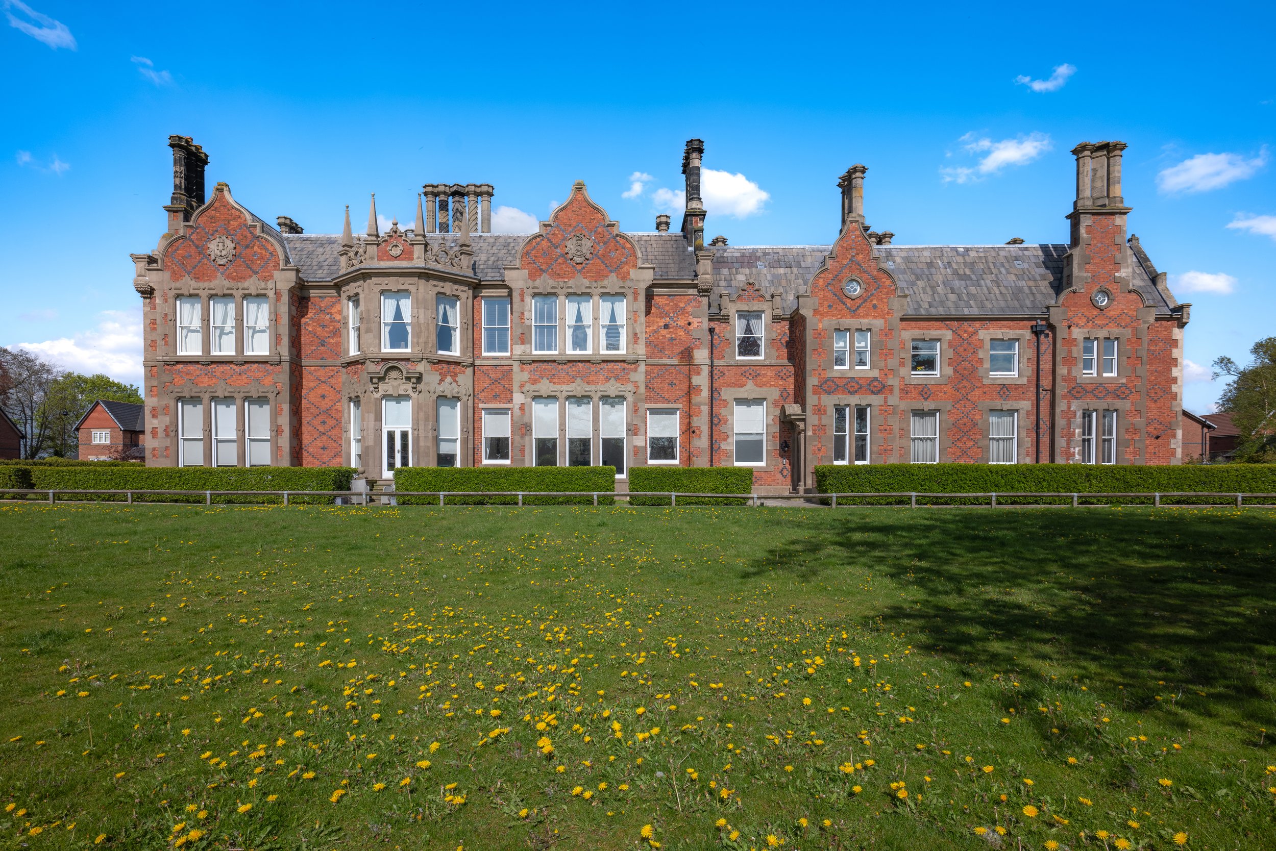 A historic brick building with ornate architectural details, surrounded by a grassy field with yellow flowers, under a bright blue sky with scattered clouds.