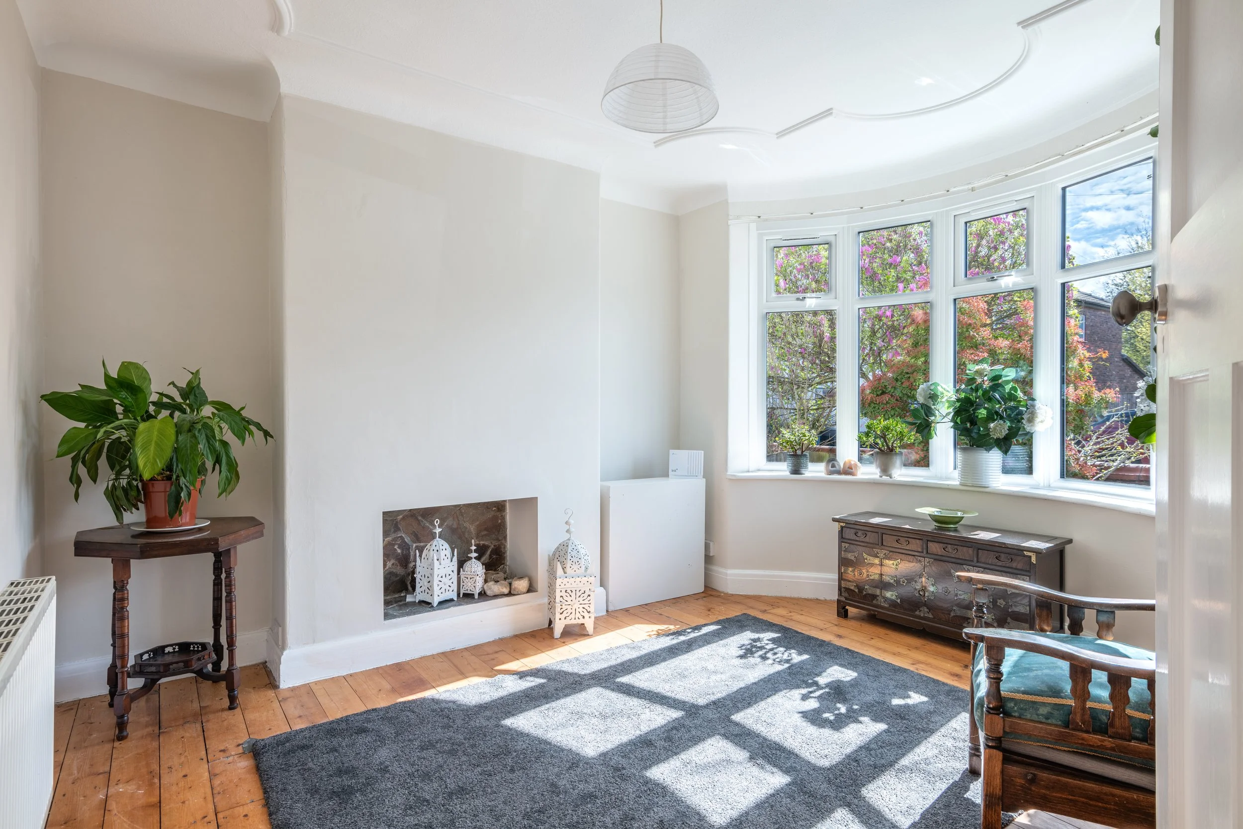 Bright living room with bay window, wooden floors, and simple decor including potted plants, a rug, and vintage furniture.